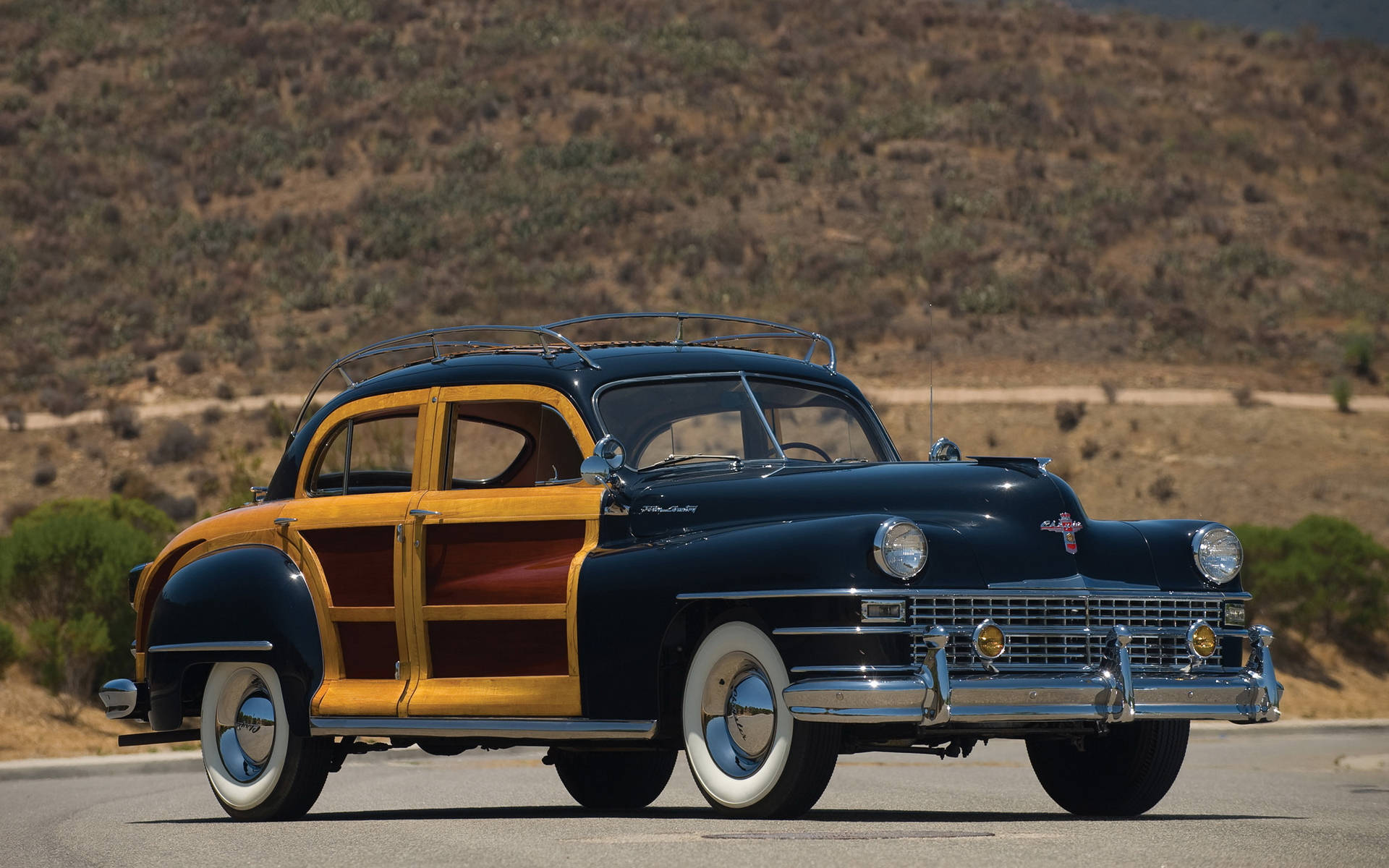 Chrysler 桌布 壁紙 — 一輛經典的1940年代木鑲板轎車在明亮的陽光下閃耀，行駛在沙漠高速公路上，背景是起伏的山丘。