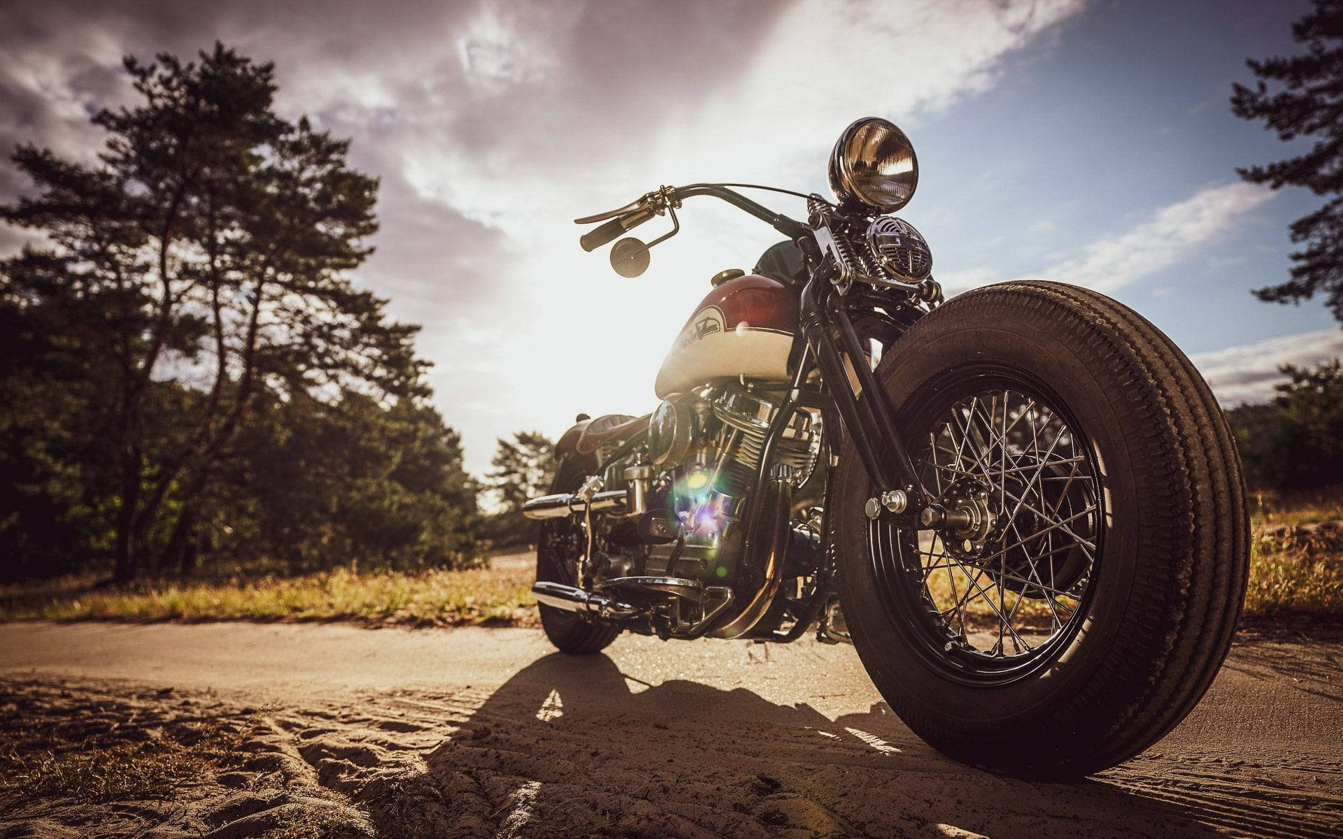 Bobber 桌布 壁紙 — 一輛經典的Chopper摩托車，帶有鍍鉻細節和紅色油箱，停在碎石路上，背景是戲劇性的日落天空和松樹。
