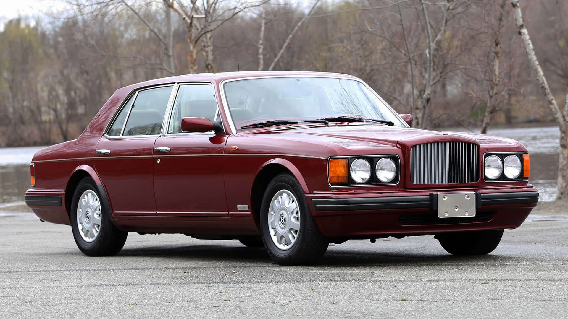 Bentley Brooklands 桌布 壁紙 — 一輛經典的栗色賓利轎車在柔和日光下閃耀，停在瀝青路面上，旁邊是一條平靜的河流，背景有光禿的樹木。