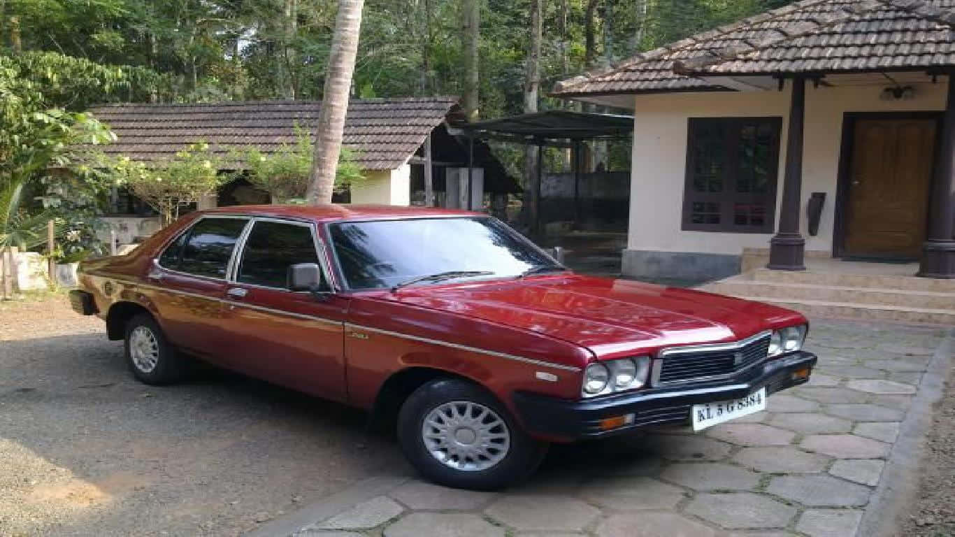 Hindustan Motors 桌布 壁紙 — 一輛經典的栗色轎車停泊在石頭車道上，旁邊是帶有瓦頂和茂密綠植的熱帶房屋。