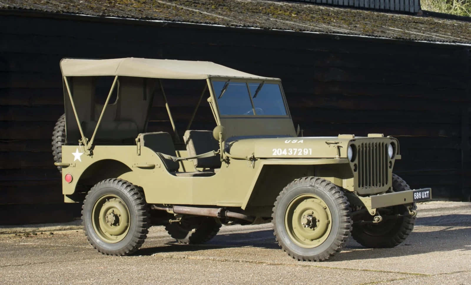 Jeep Willys 桌布 壁紙 — 一輛經典的橄欖綠軍用吉普車，帶帆布頂篷，停在碎石路上，前面是深色木製穀倉，在明亮的日光下。