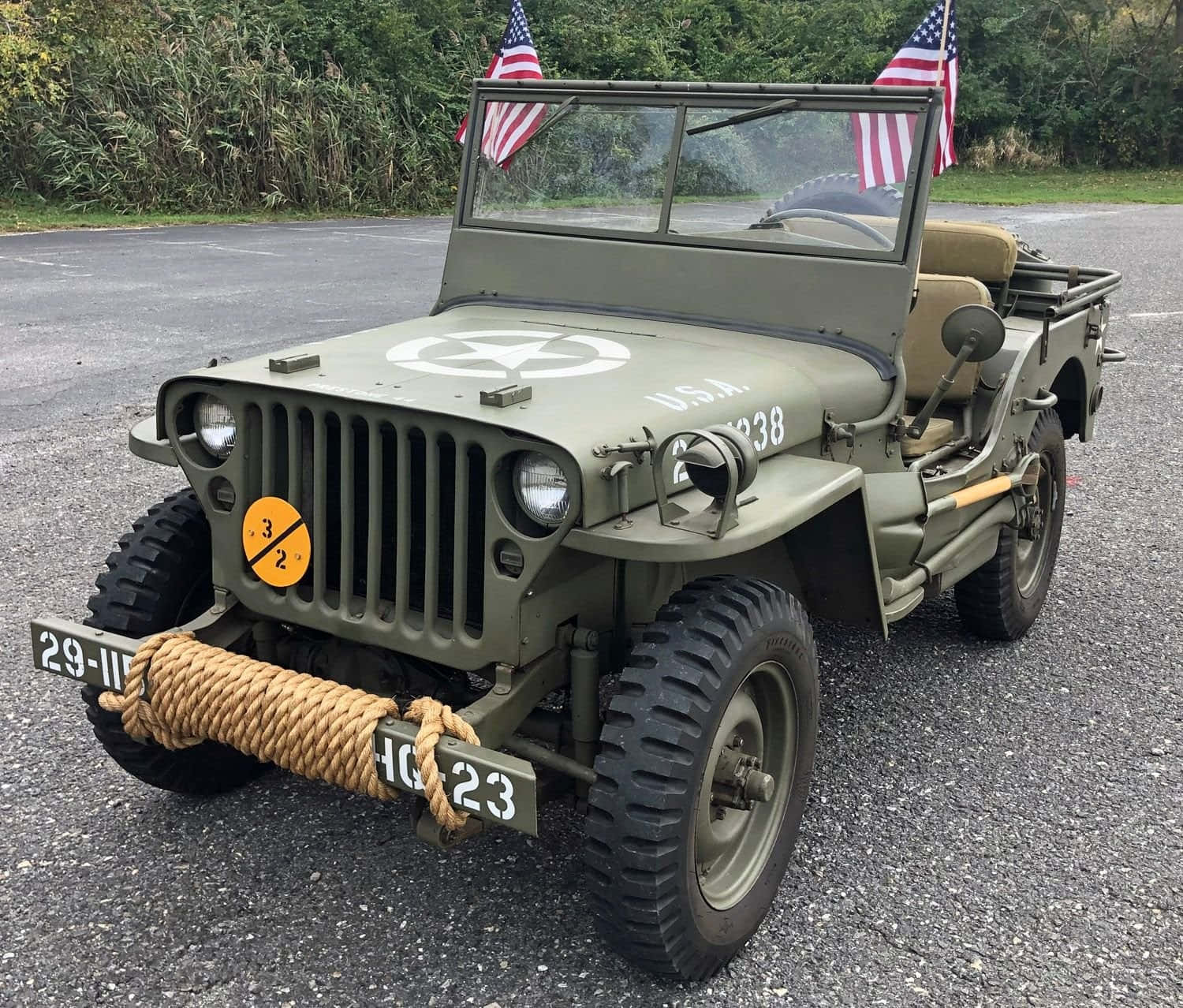 Jeep Willys 桌布 壁紙 — 一輛經典的橄欖綠軍用吉普車，帶有美國國旗和繩索保險桿，停在柏油路上，靠近綠色植物。