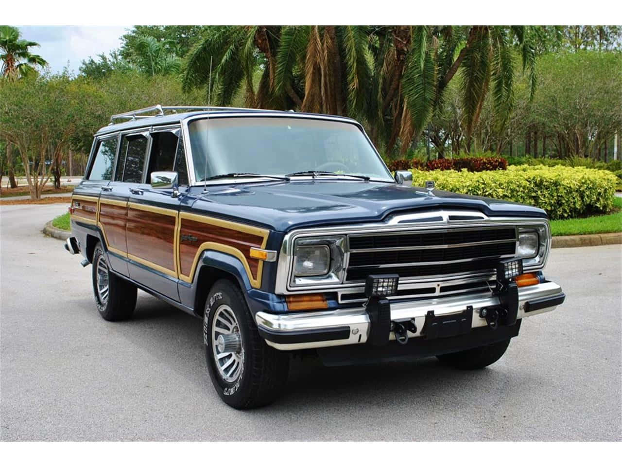 Jeep Wagoneer 桌布 壁紙 — 一輛經典的藍色木紋Jeep Wagoneer，帶有鍍鉻裝飾，停在鋪砌的道路上，周圍環繞著棕櫚樹和修剪整齊的灌木。