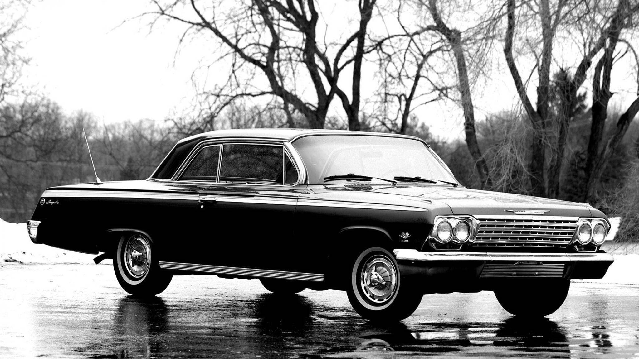 chevrolet impala 1967 桌布 壁紙 — 一輛經典的黑色1960年代雪佛蘭Impala在濕漉漉的道路上閃閃發光，背景是光禿禿的冬季樹木，這是一張清晰的黑白照片。