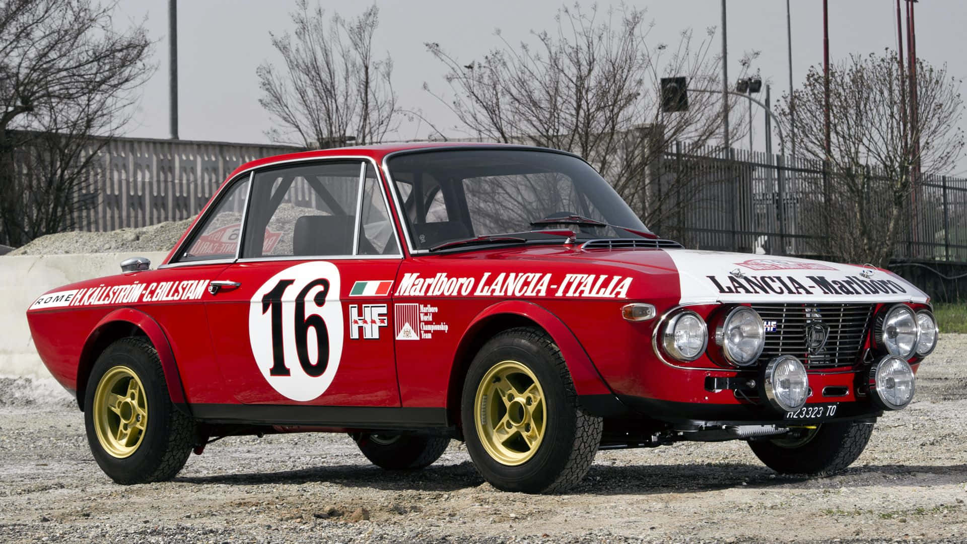 Lancia 桌布 壁紙 — 一輛經典紅白相間的藍旗亞拉力賽車，編號16，停泊在碎石地上，附近有光禿禿的樹木。