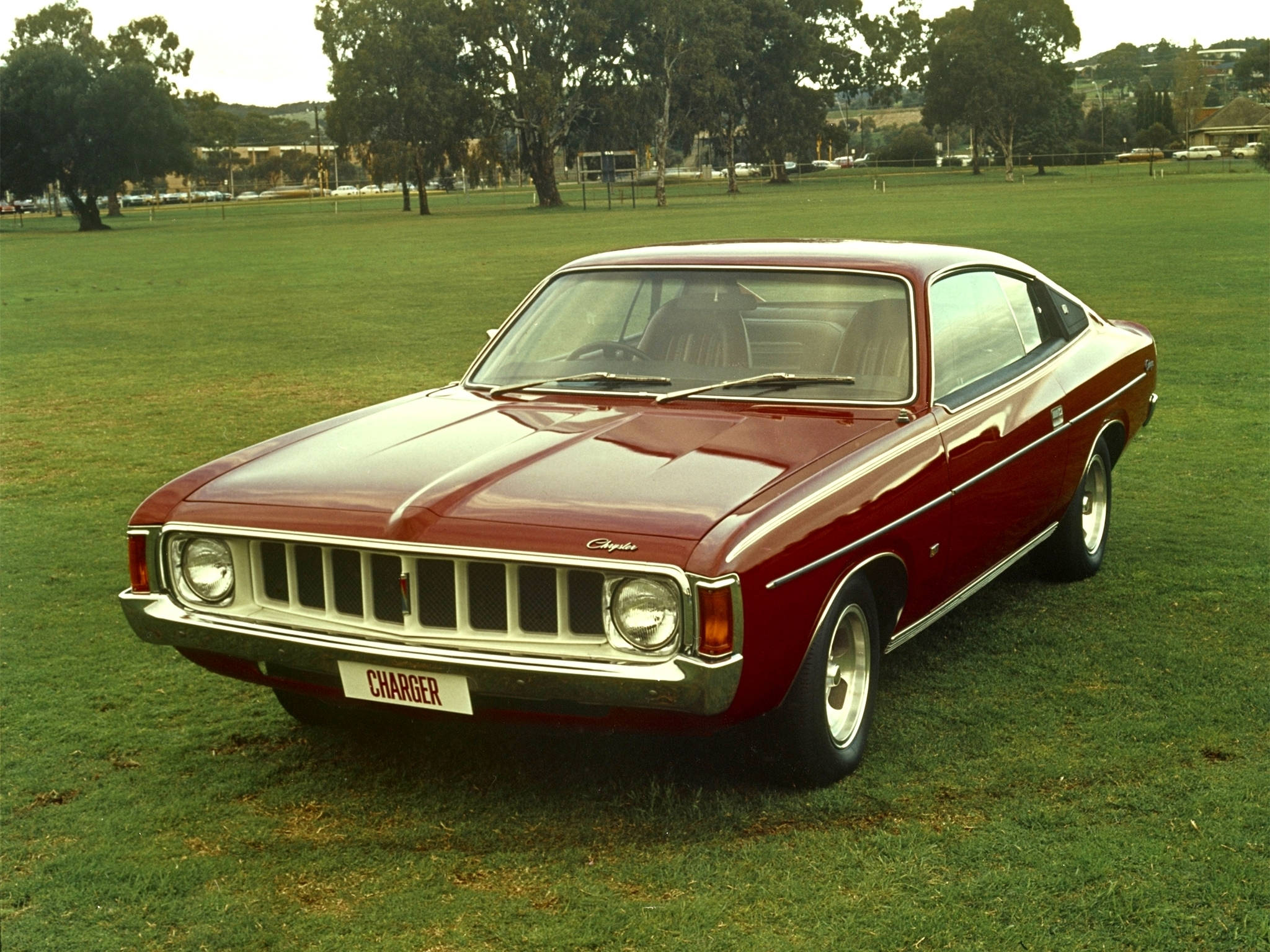 Chrysler 桌布 壁紙 — 一輛經典紅色Chrysler Charger雙門跑車停放在茂密的綠色草坪上，背景有樹木和遠處建築物，在柔和日光下。