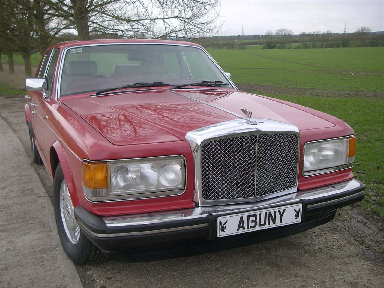 Bentley Eight 桌布 壁紙 — 一輛經典紅色賓利 Turbo R 停泊在碎石路上，旁邊是綠色的田野，天空多雲。
