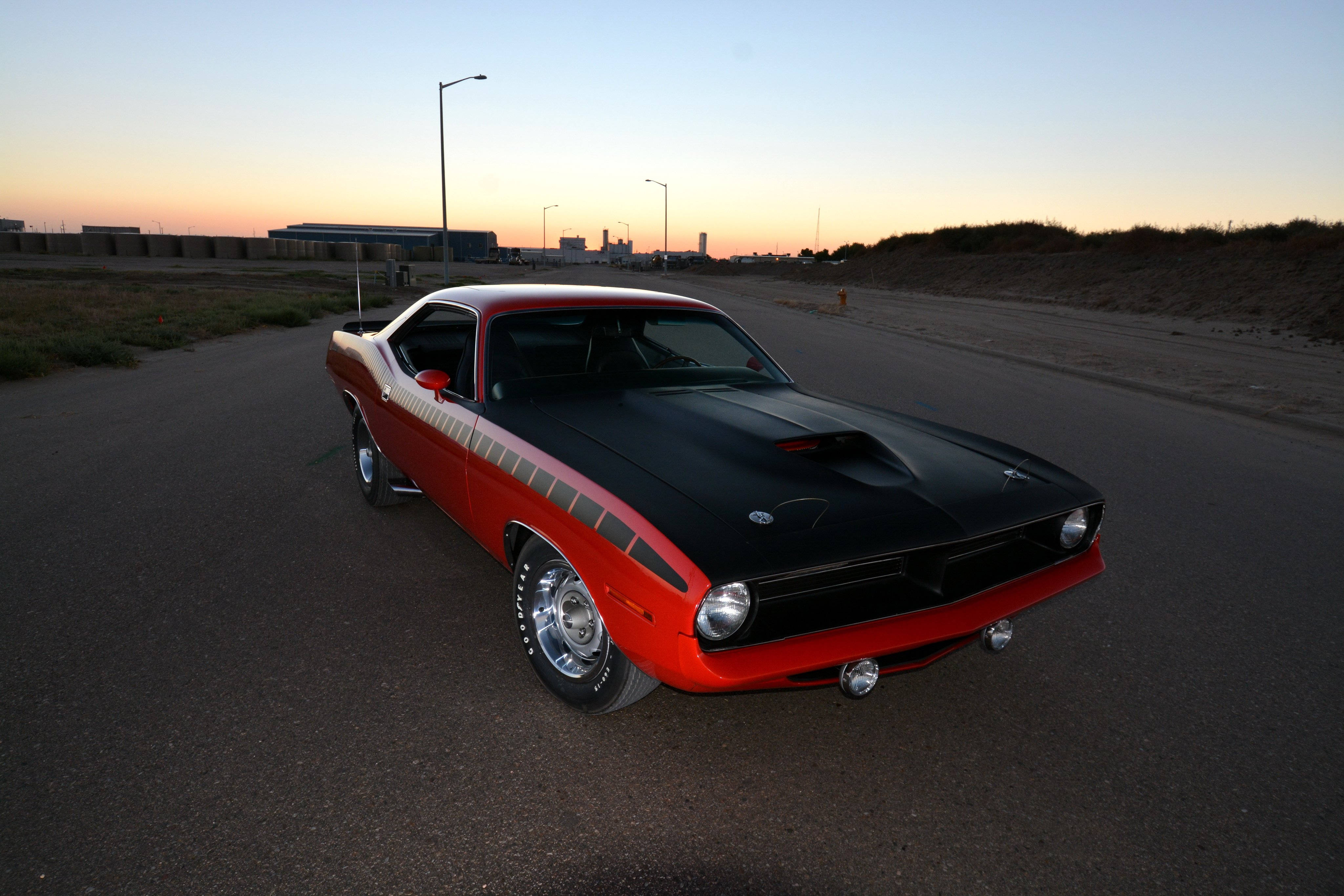 Barracuda 桌布 壁紙 — 一輛經典紅黑色肌肉車在日落時分停泊在空曠的道路上，遠處有工業建築。