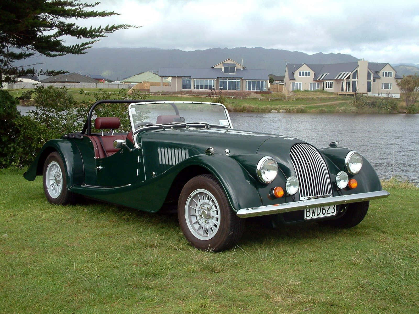 Morgan 桌布 壁紙 — 一輛經典綠色摩根敞篷跑車，配紅色皮革座椅，停在湖邊草地上，背景有房屋和山丘，天空多雲。