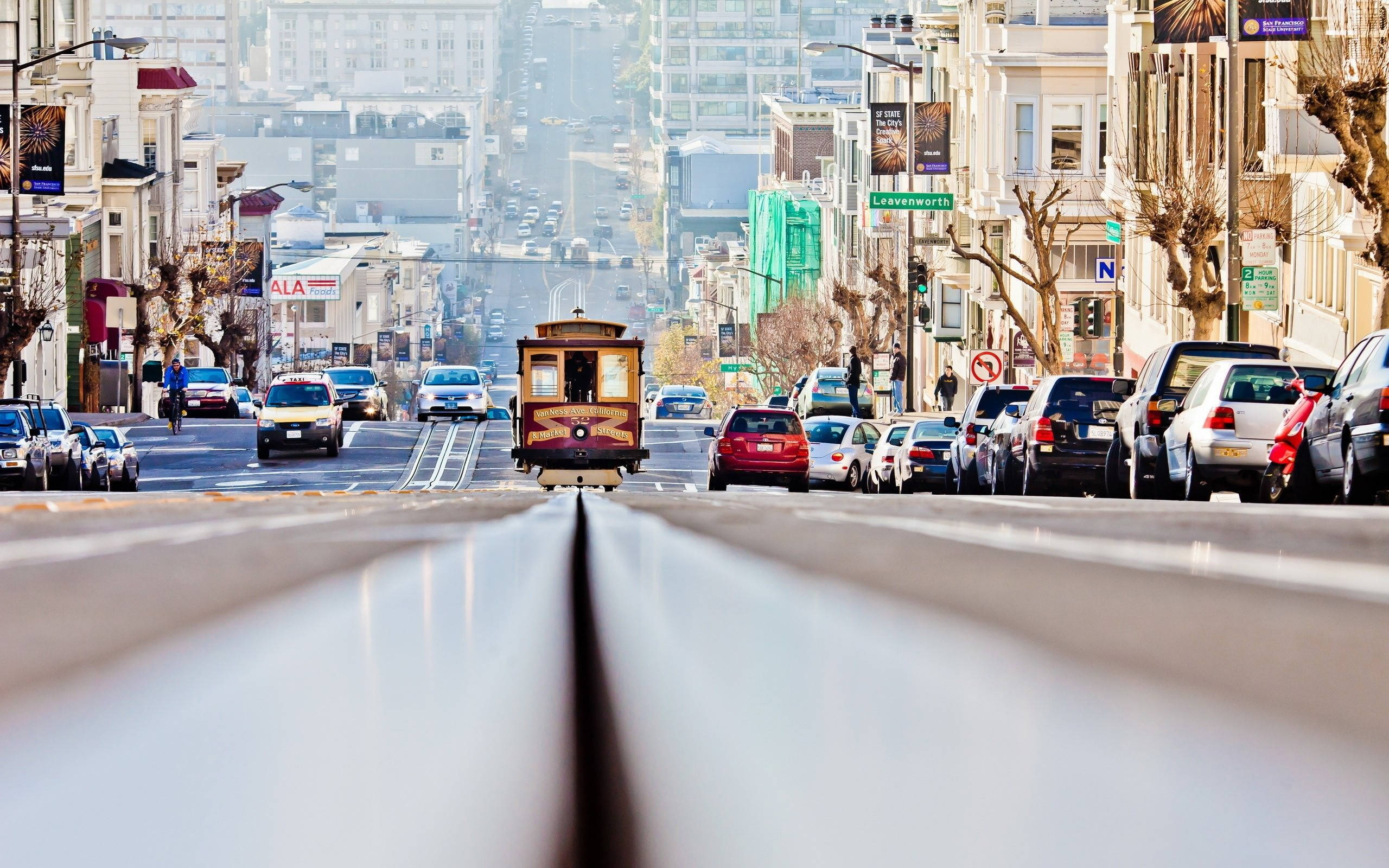 City Street 桌布 壁紙 — 一輛經典纜車在明亮的日光下，沿著舊金山一條陡峭的街道上行，街道兩旁停滿了汽車，樹木光禿。