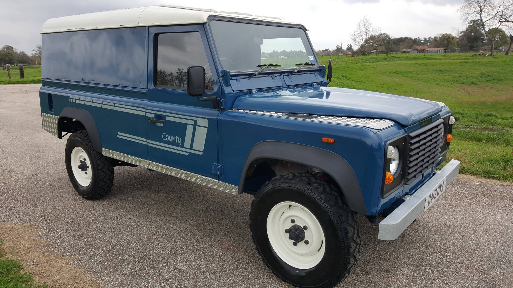land rover iPhone 桌布 壁紙 — 一輛經典藍色 Land Rover Defender County 廂型車停泊在碎石路上，旁邊是草地，天空多雲。