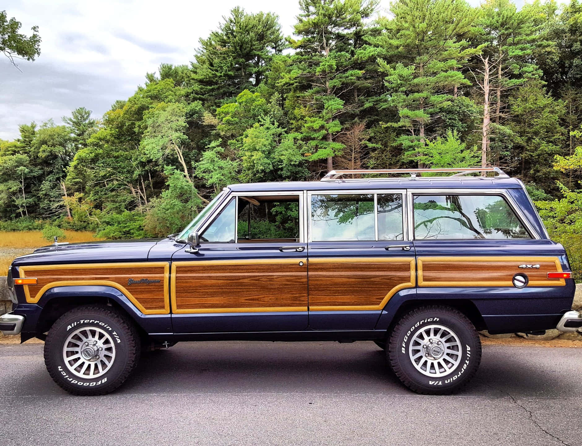 Jeep Wagoneer 桌布 壁紙 — 一輛經典藍色木紋車身Jeep Cherokee XJ配銀色輪圈，停泊在陰天下一條鋪砌道路旁，旁邊是茂密的綠色森林。