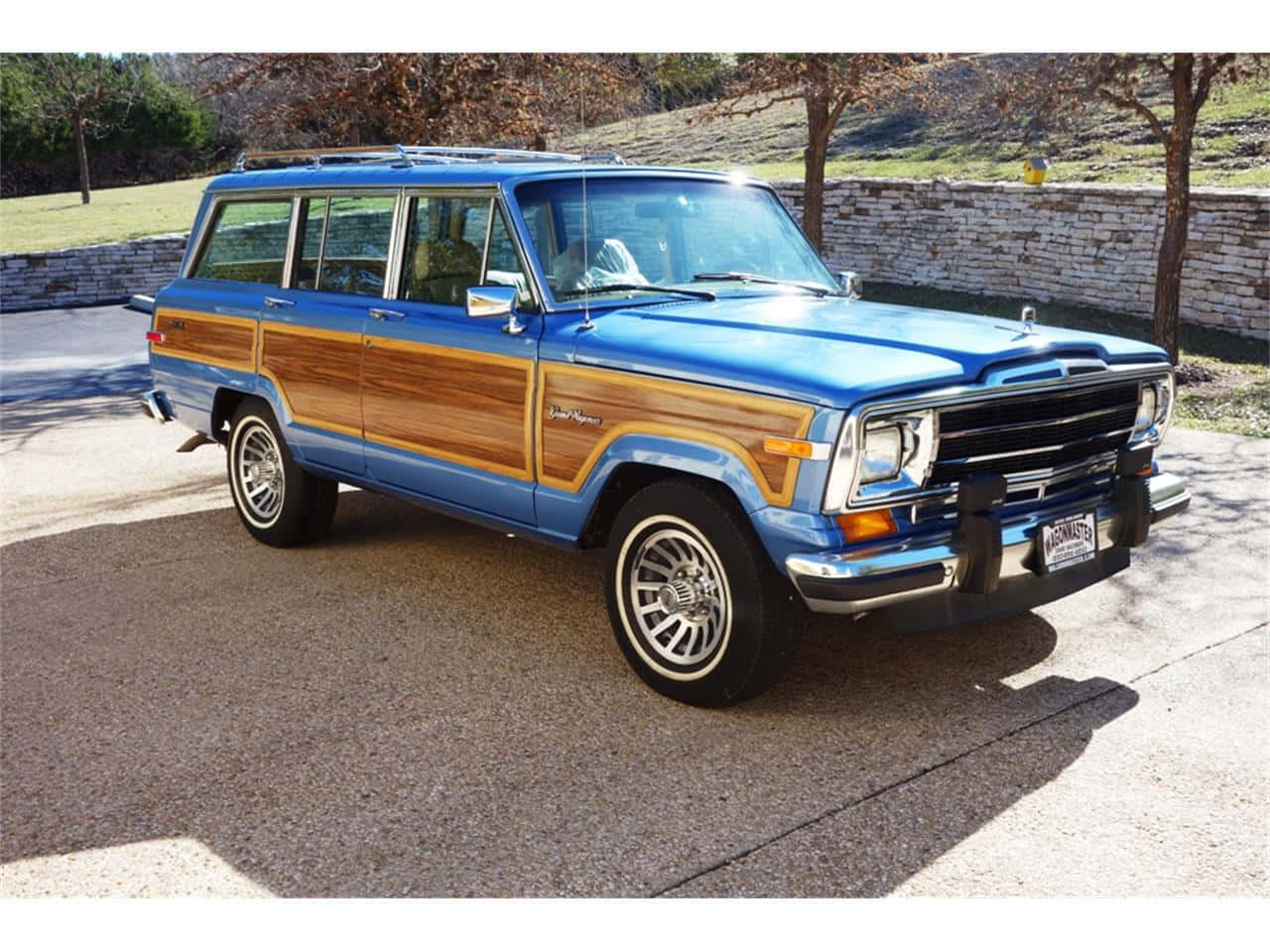 Jeep Wagoneer 桌布 壁紙 — 一輛經典藍色木紋車身Jeep Wagoneer停放在陽光普照的混凝土車道上，旁邊有石牆和樹木。