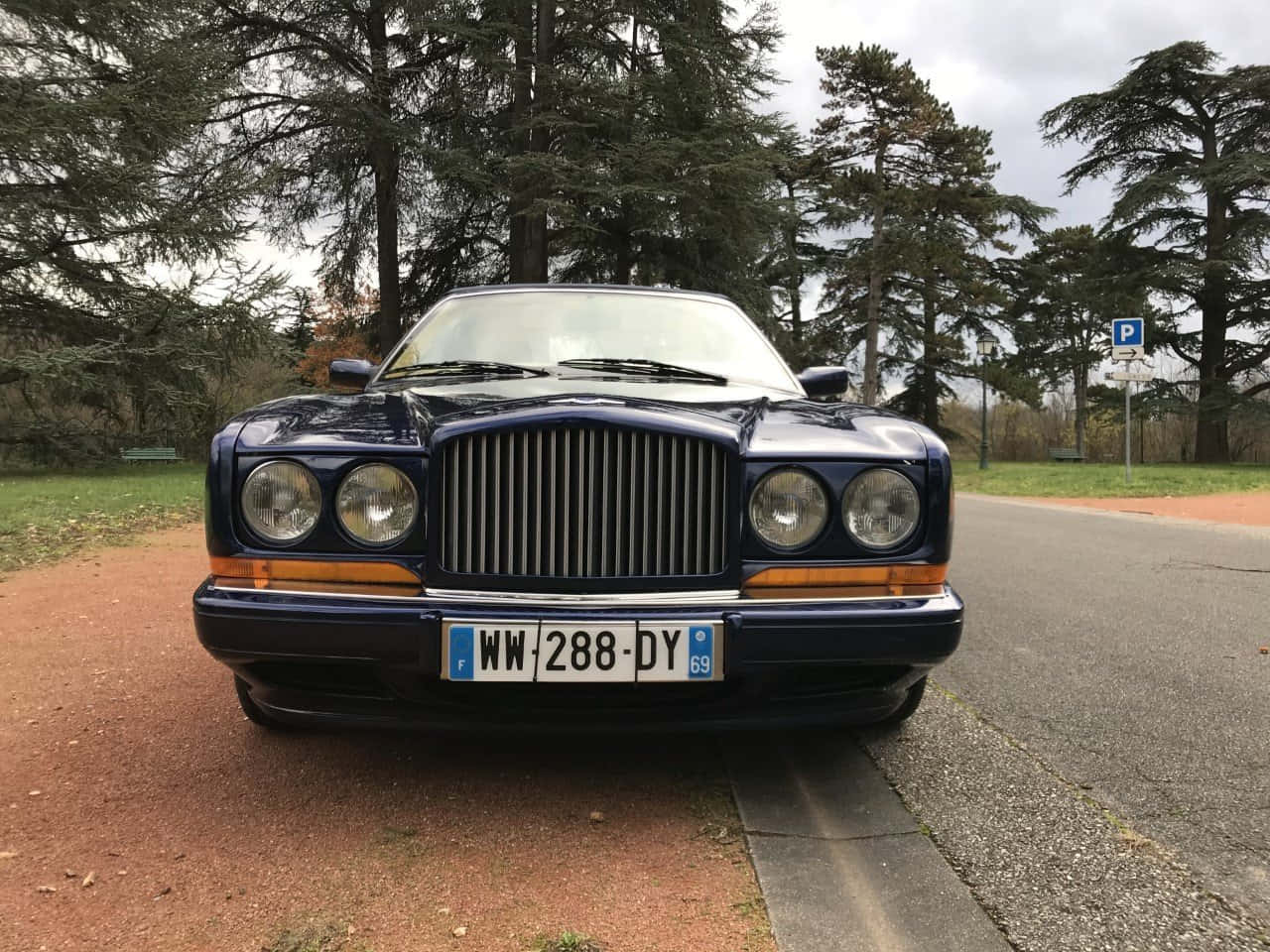 Bentley Azure 桌布 壁紙 — 一輛經典藍色賓利停泊在樹蔭大道旁的碎石路上，天空多雲。