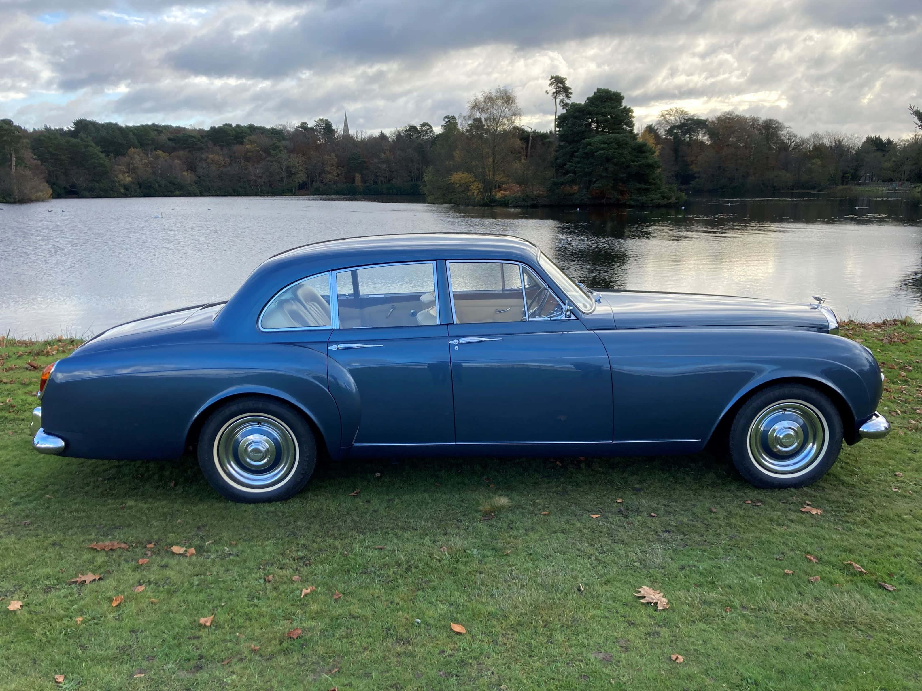 Bentley S3 桌布 壁紙 — 一輛經典藍色賓利房車停泊在平靜湖邊的草地上，天空多雲。