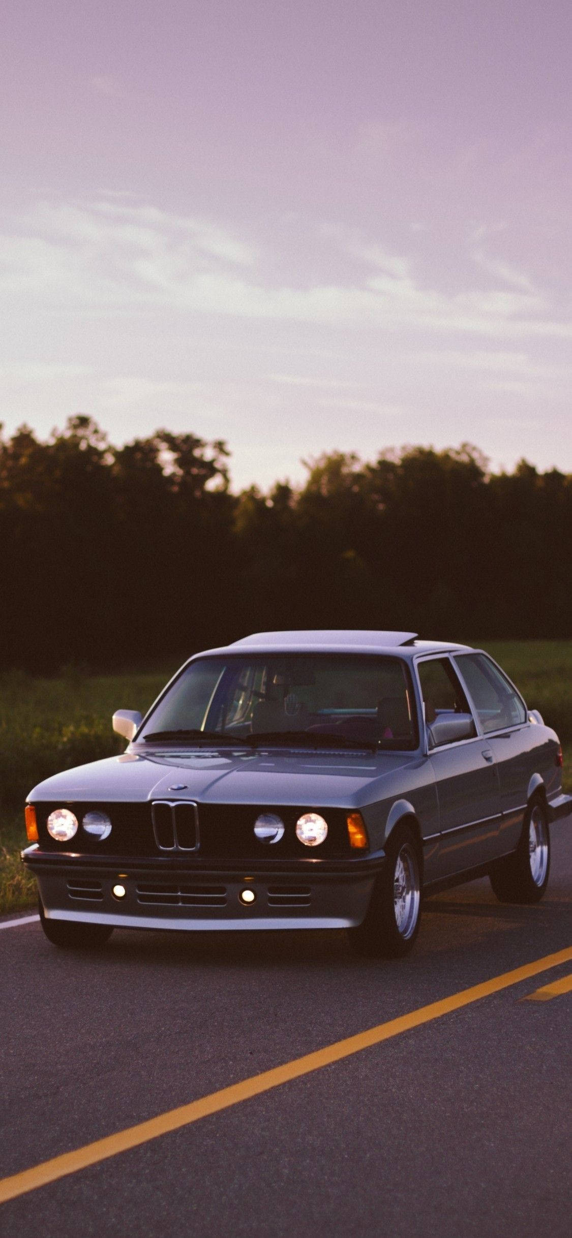 X 桌布 壁紙 — 一輛經典銀色BMW E30轎車在日落時分沿鄉村道路滑行，溫暖的金色光芒沐浴其流暢線條和閃爍的車頭燈。