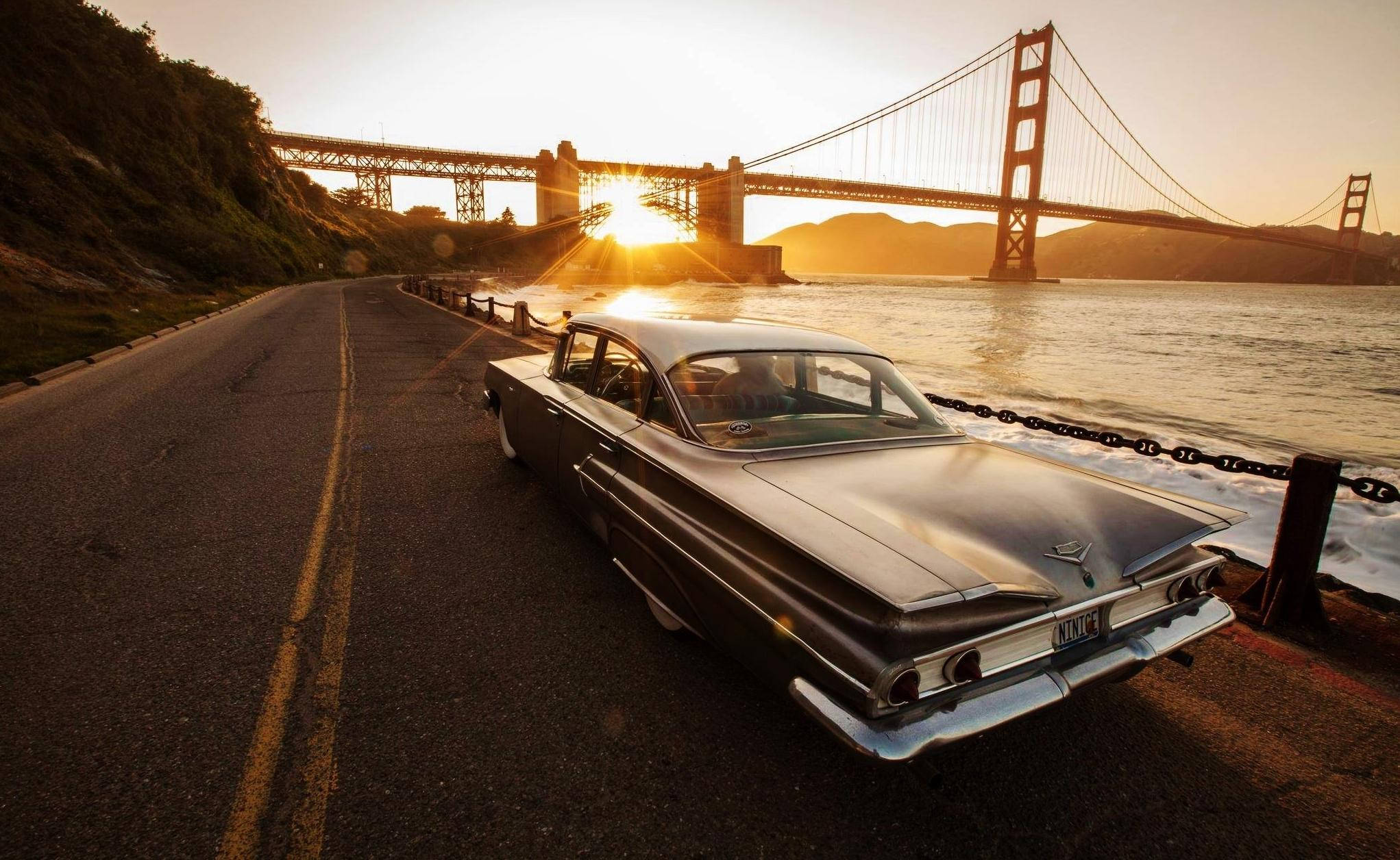 Cadillac 桌布 壁紙 — 一輛經典銀色汽車停泊在海濱公路上，背後的金門大橋在夕陽餘暉中閃耀。