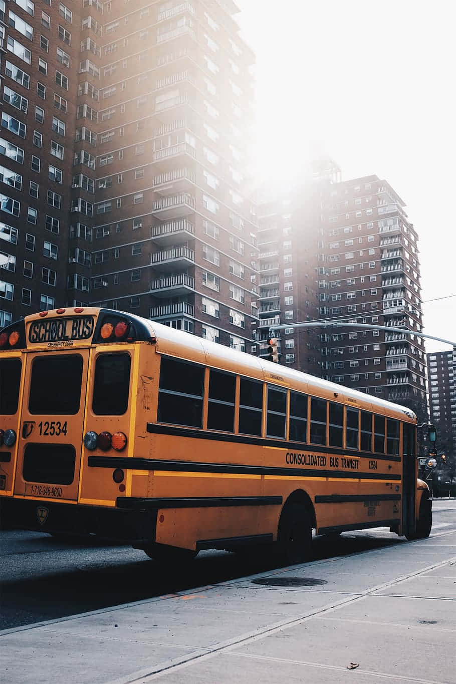 Bus 桌布 壁紙 — 一輛經典黃色校車停泊在城市的街道上，背後有高聳的磚砌公寓大樓，在明亮的陽光下顯得格外醒目。