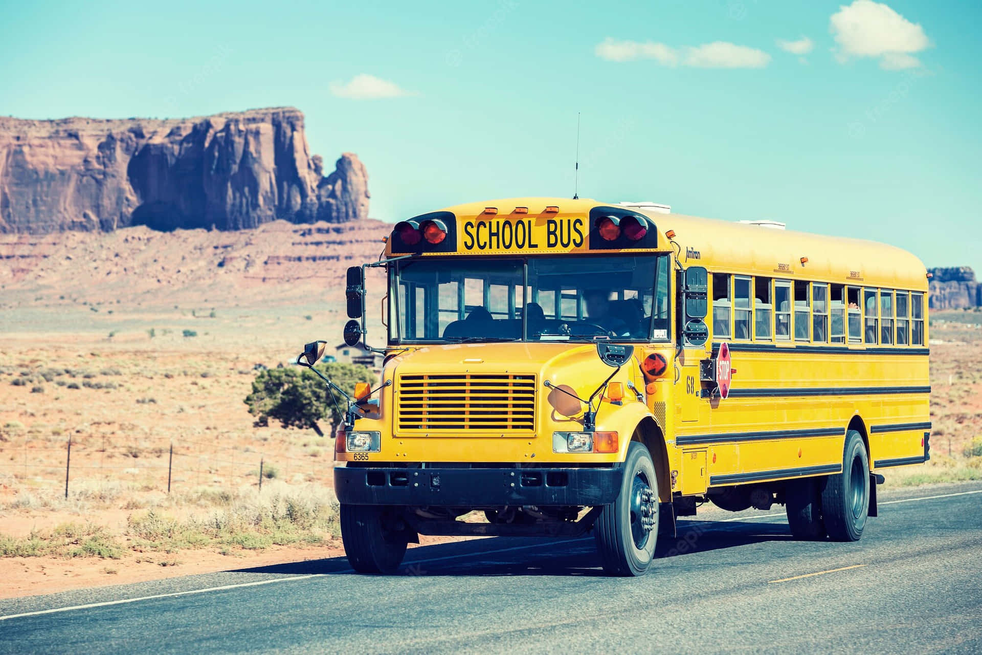 Yellow School Bus 桌布 壁紙 — 一輛經典黃色校車行駛在沙漠公路上，周圍是紅色岩石地貌，上方是明亮的藍天。