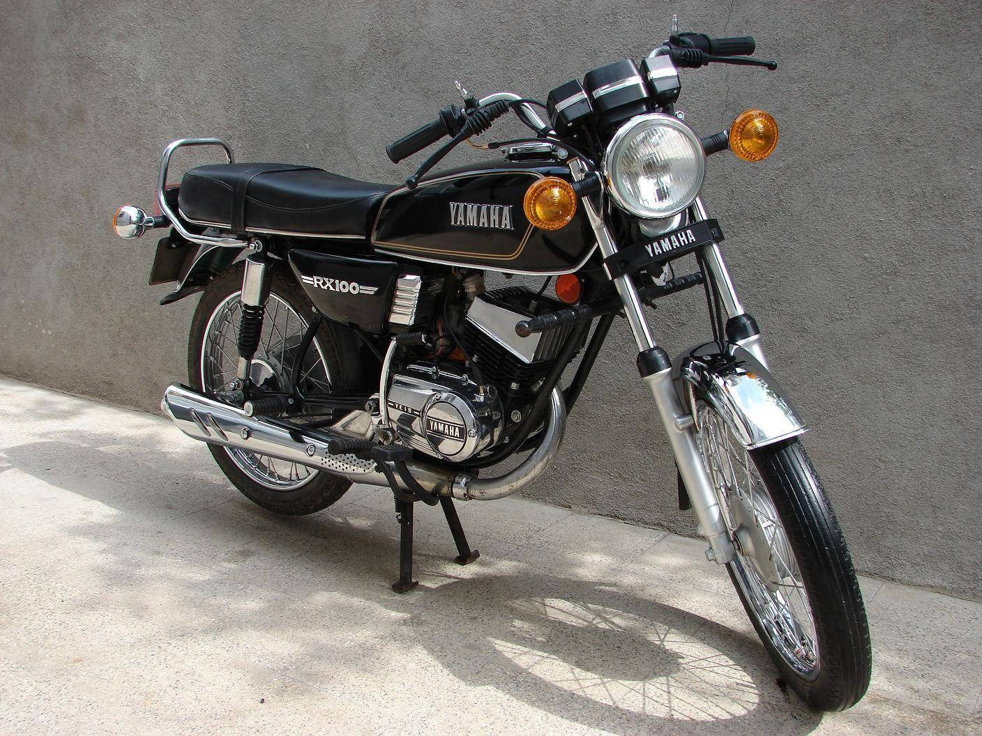 Rx100 桌布 壁紙 — 一輛經典黑色Yamaha RX100摩托車停放在混凝土上，背景是一面素色灰牆，在明亮的日光下。