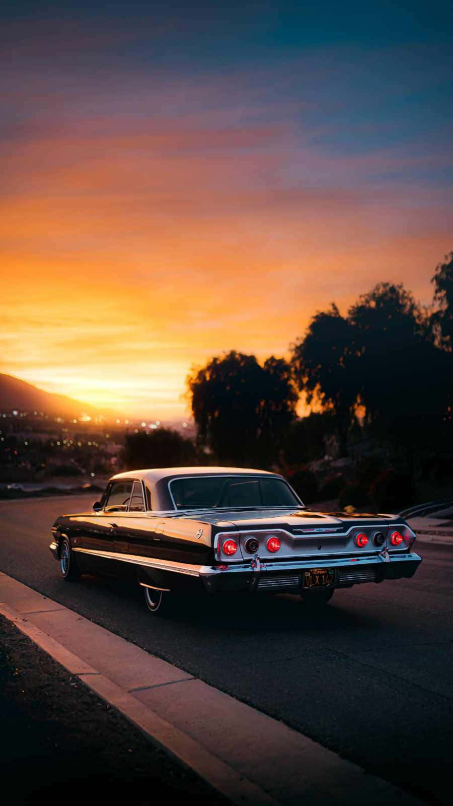 Vintage Car iPhone 桌布 壁紙 — 一輛經典黑色和銀色汽車，帶有發光的紅色尾燈，停在日落時分的街道上，天空呈現橙色和藍色。
