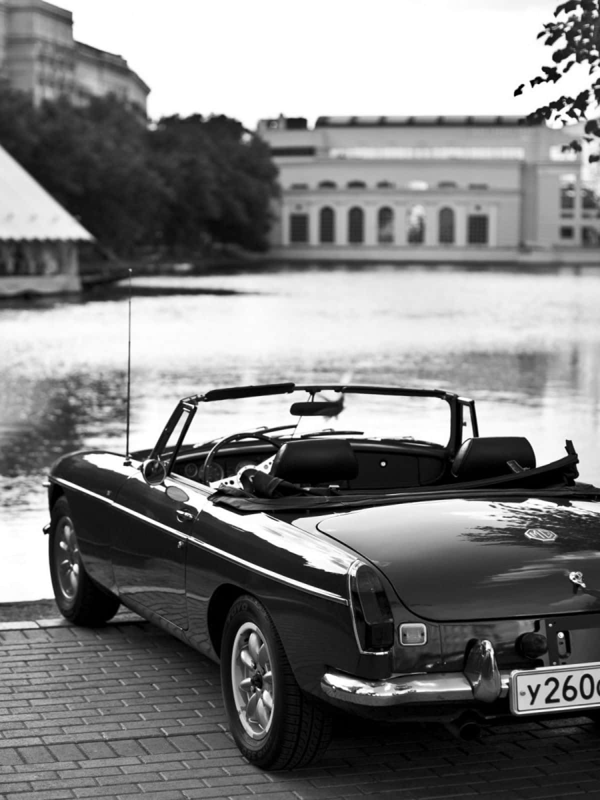 Black And White Car 桌布 壁紙 — 一輛經典黑色敞篷車停泊在磚砌碼頭旁，旁邊是平靜的水面，背景是歷史建築。