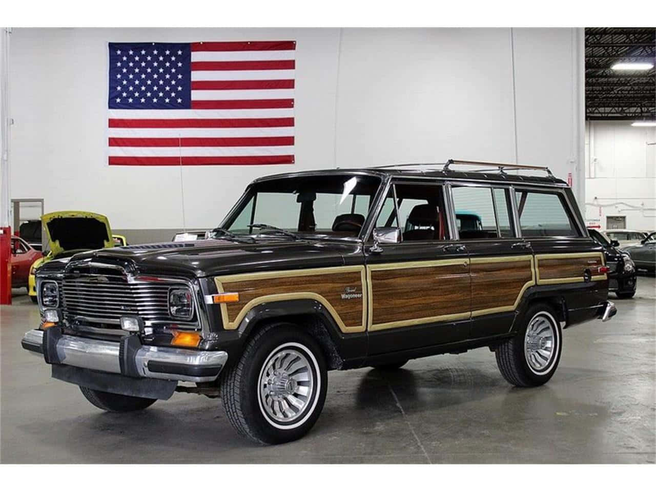 Jeep Wagoneer 桌布 壁紙 — 一輛經典黑色木紋車身、配備鉻合金輪圈的Jeep Wagoneer，在美國國旗下自豪地停放在車庫中。