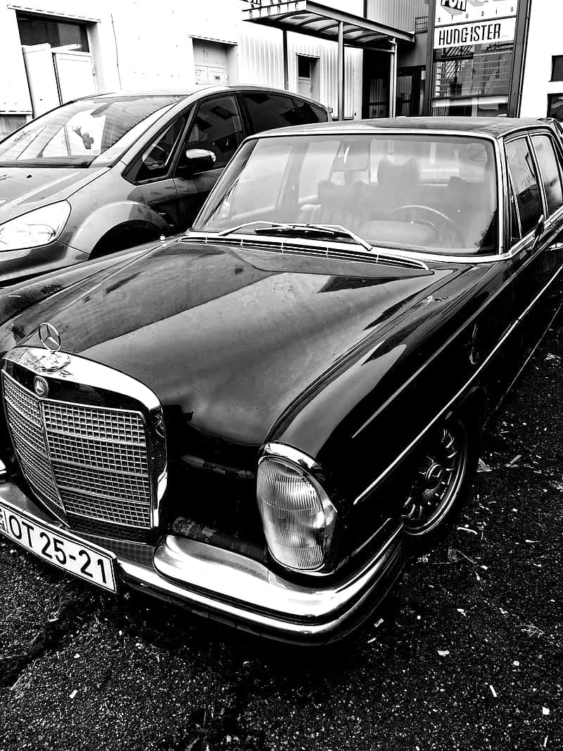 Classic Mercedes 桌布 壁紙 — 一輛經典黑色賓士轎車在高對比度的黑白中閃耀，停放在現代廂型車旁邊的市區停車場。