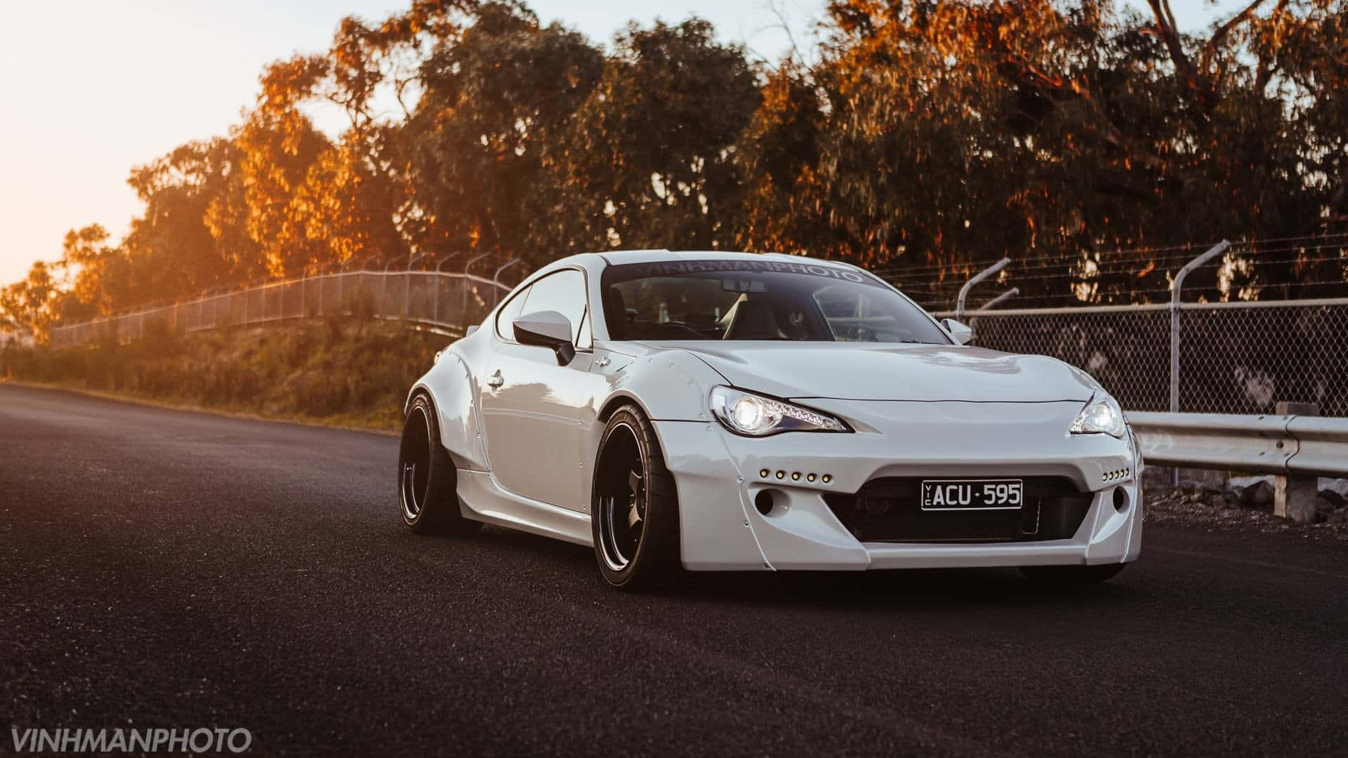 日落時分，白色改裝跑車行駛在路上，背景有樹木和圍欄。 — Toyota Supra Mk4 壁紙