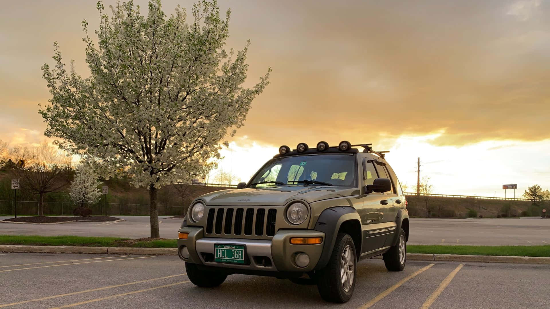 Jeep Liberty 桌布 壁紙 — 一輛綠色Jeep Liberty停放在停車場，旁邊有一棵盛開的樹，背景是金色的日落天空。
