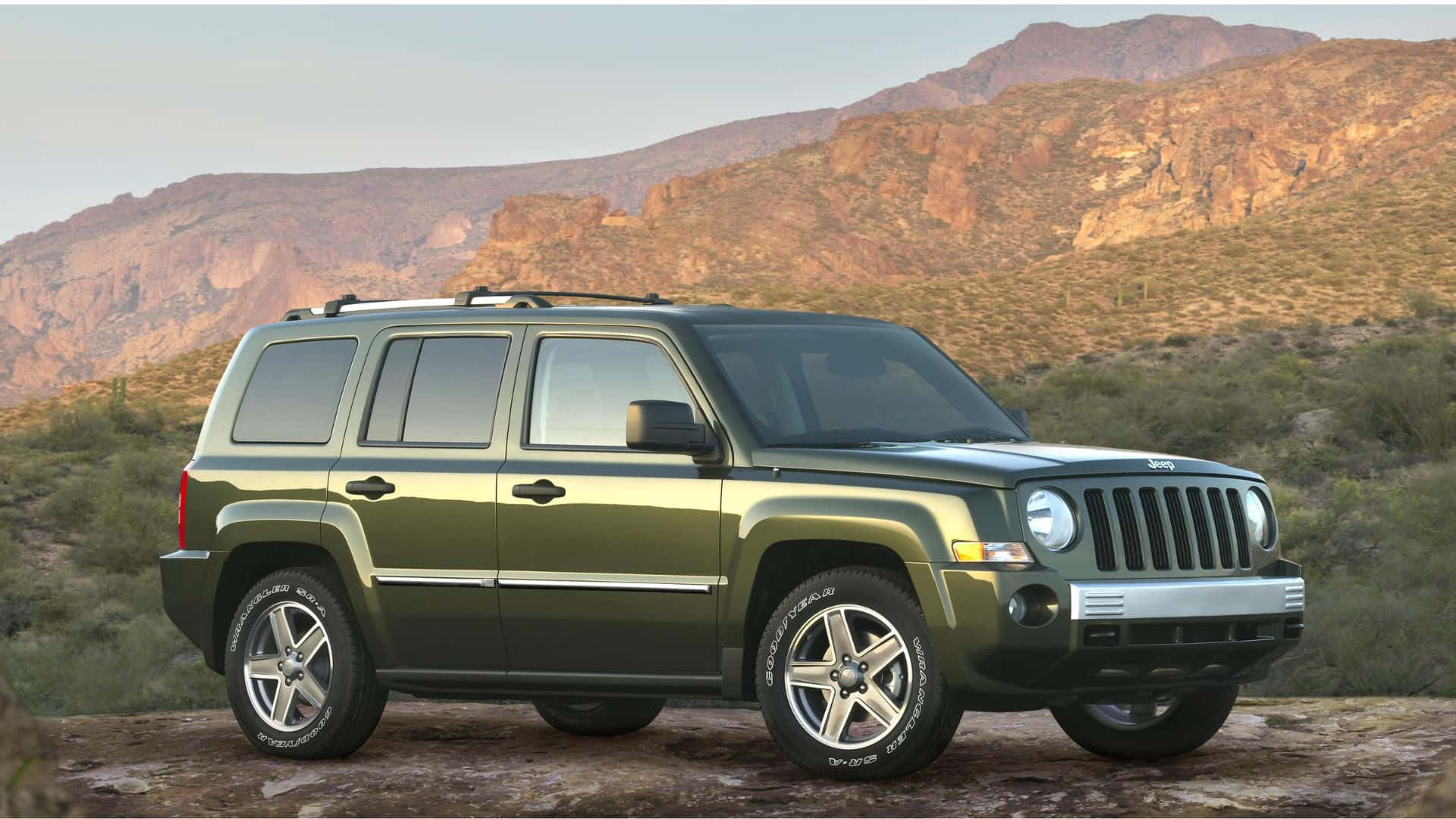 Jeep Patriot 桌布 壁紙 — 一輛綠色Jeep Patriot SUV在岩石沙漠地形上，背景是崎嶇的山脈和稀疏的仙人掌，天空呈現柔和的金色。