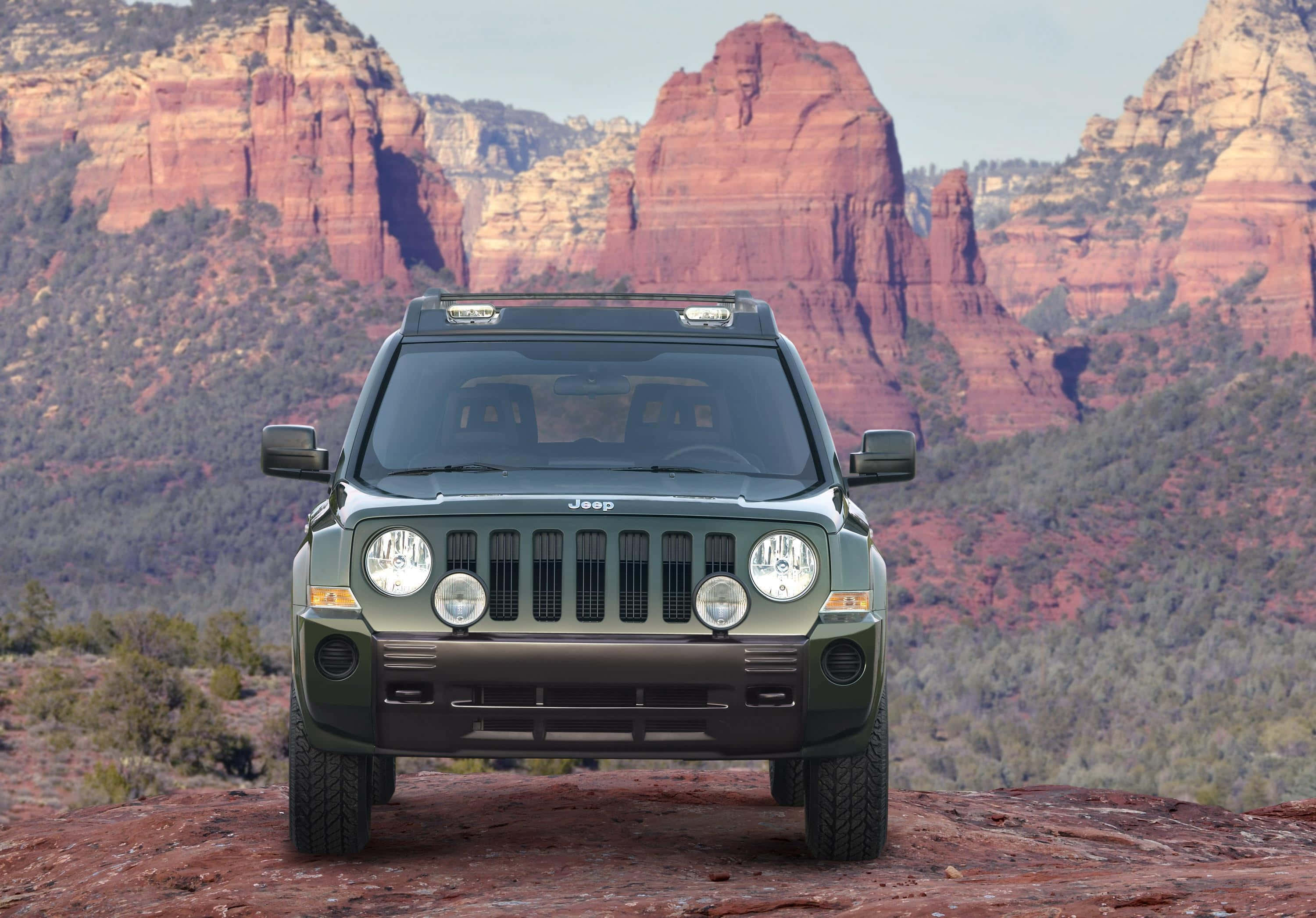 Jeep Patriot 桌布 壁紙 — 一輛綠色Jeep Wrangler在晴朗天空下，雄偉地矗立在紅色岩石地形上，背後是高聳的深紅色峽谷牆壁。