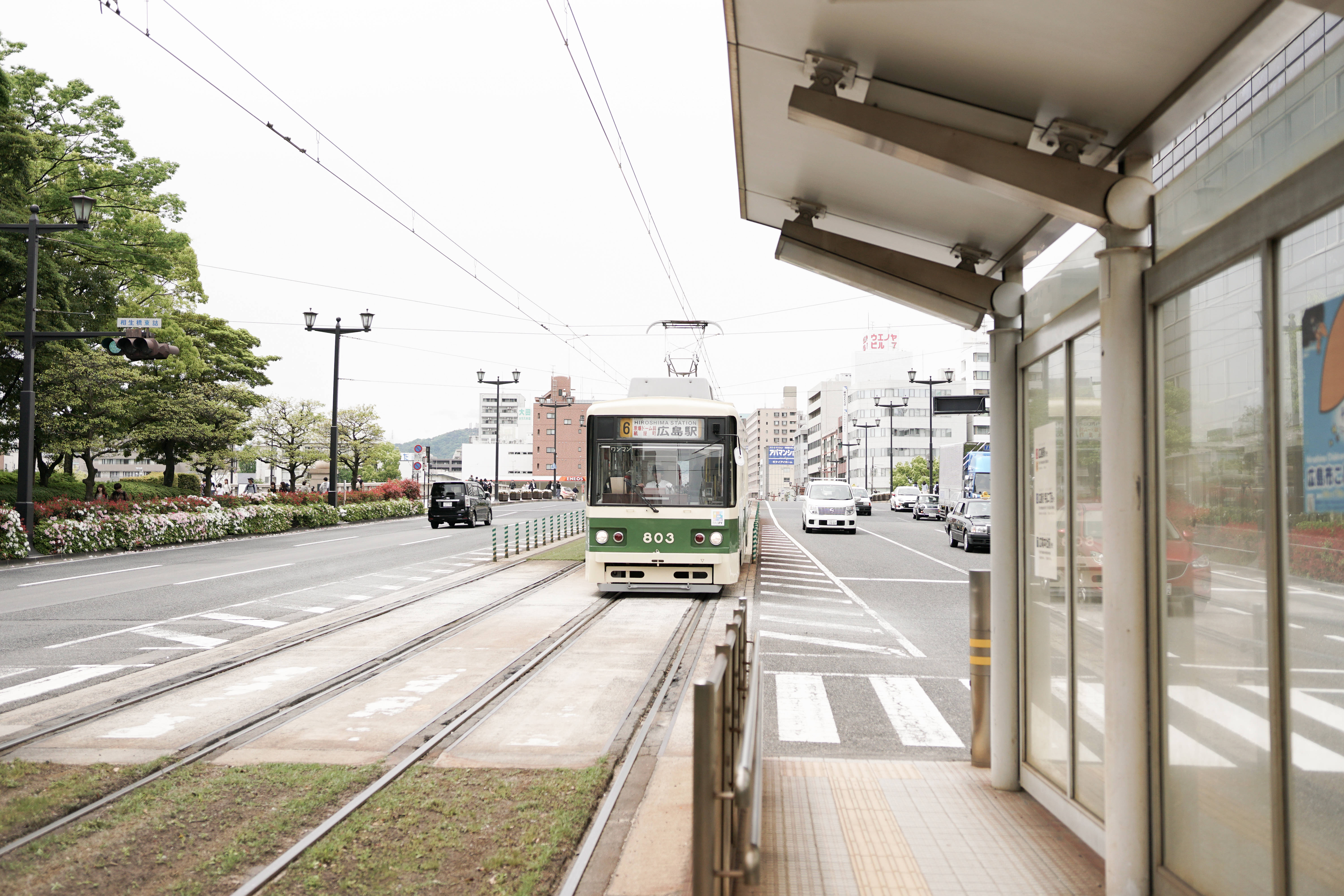火車 桌布 壁紙 — 一輛綠色和白色的電車在晴朗多雲的日子裡，沿著兩旁種有樹木和建築物的街道，接近一個現代化的車站月台。
