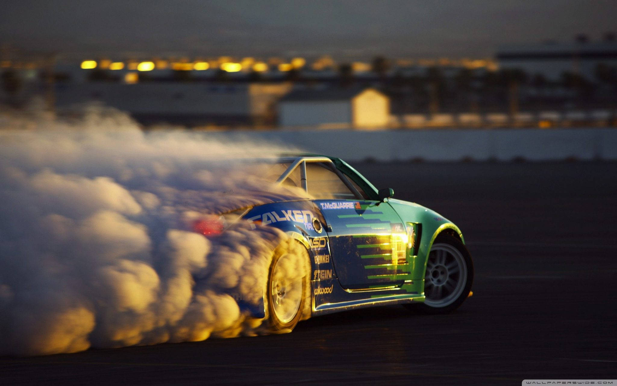 drifting 桌布 壁紙 — 一輛綠色和藍色的飄移車在黃昏時分在黑暗的瀝青賽道上滑行，揚起濃煙，背景中城市燈光閃爍。