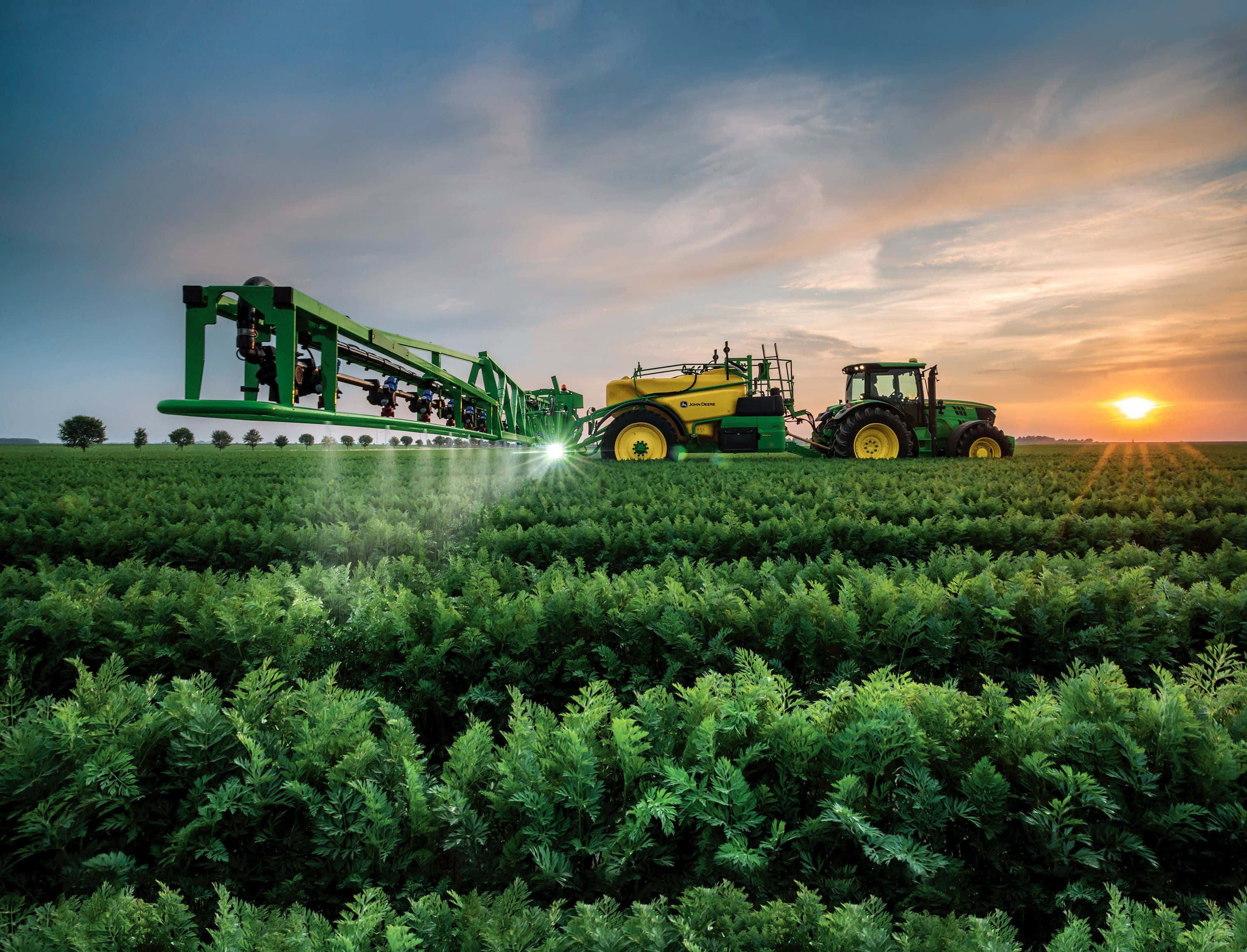 Agriculture 桌布 壁紙 — 一輛綠色和黃色的拖拉機在日落時分拖著噴霧器穿過廣闊的翠綠農田。