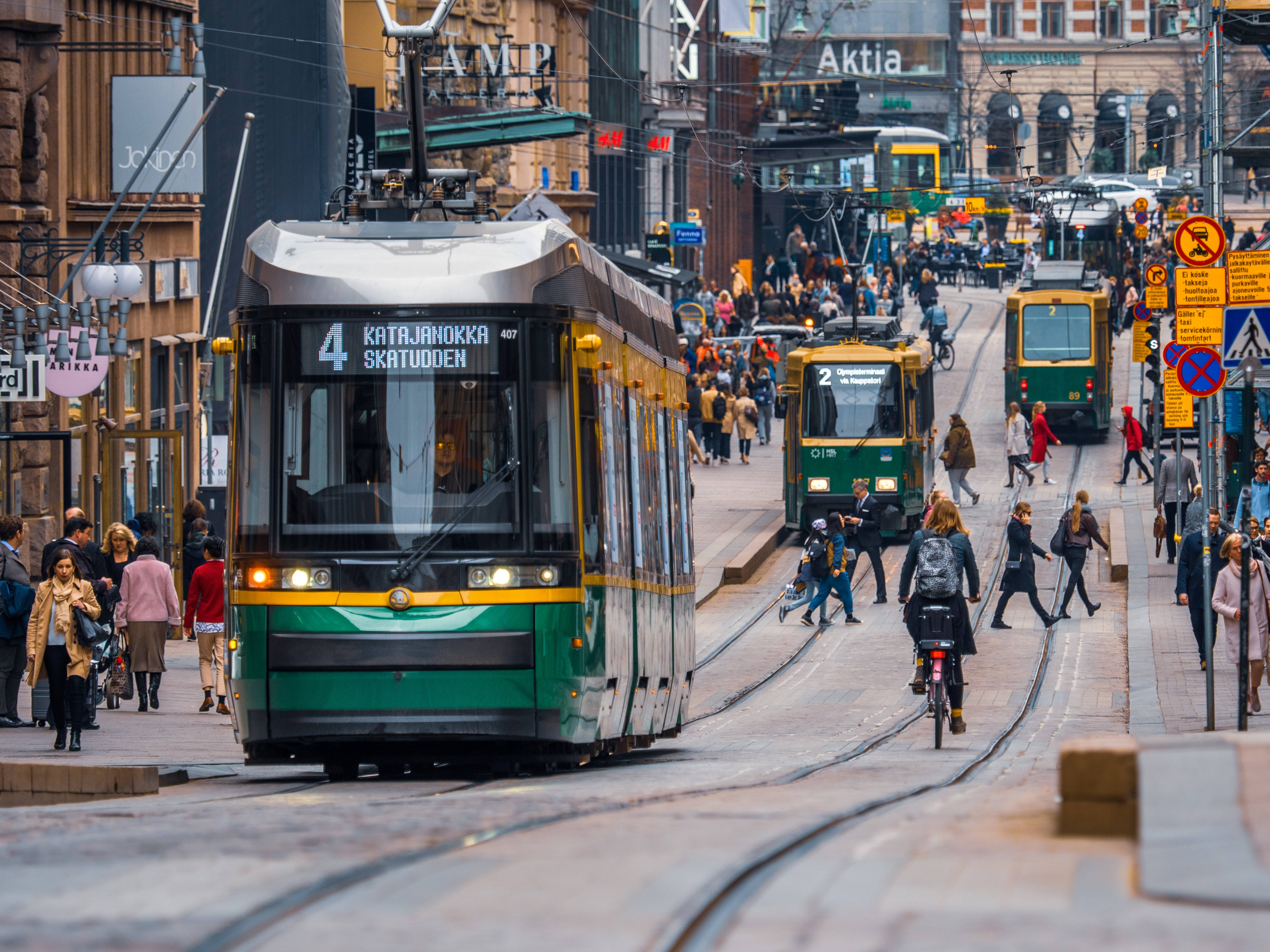 Finland 桌布 壁紙 — 一輛綠色和黃色的電車在明亮的日光下行駛在一條繁忙的城市街道上，街道上滿是行人、騎自行車者和歷史建築。