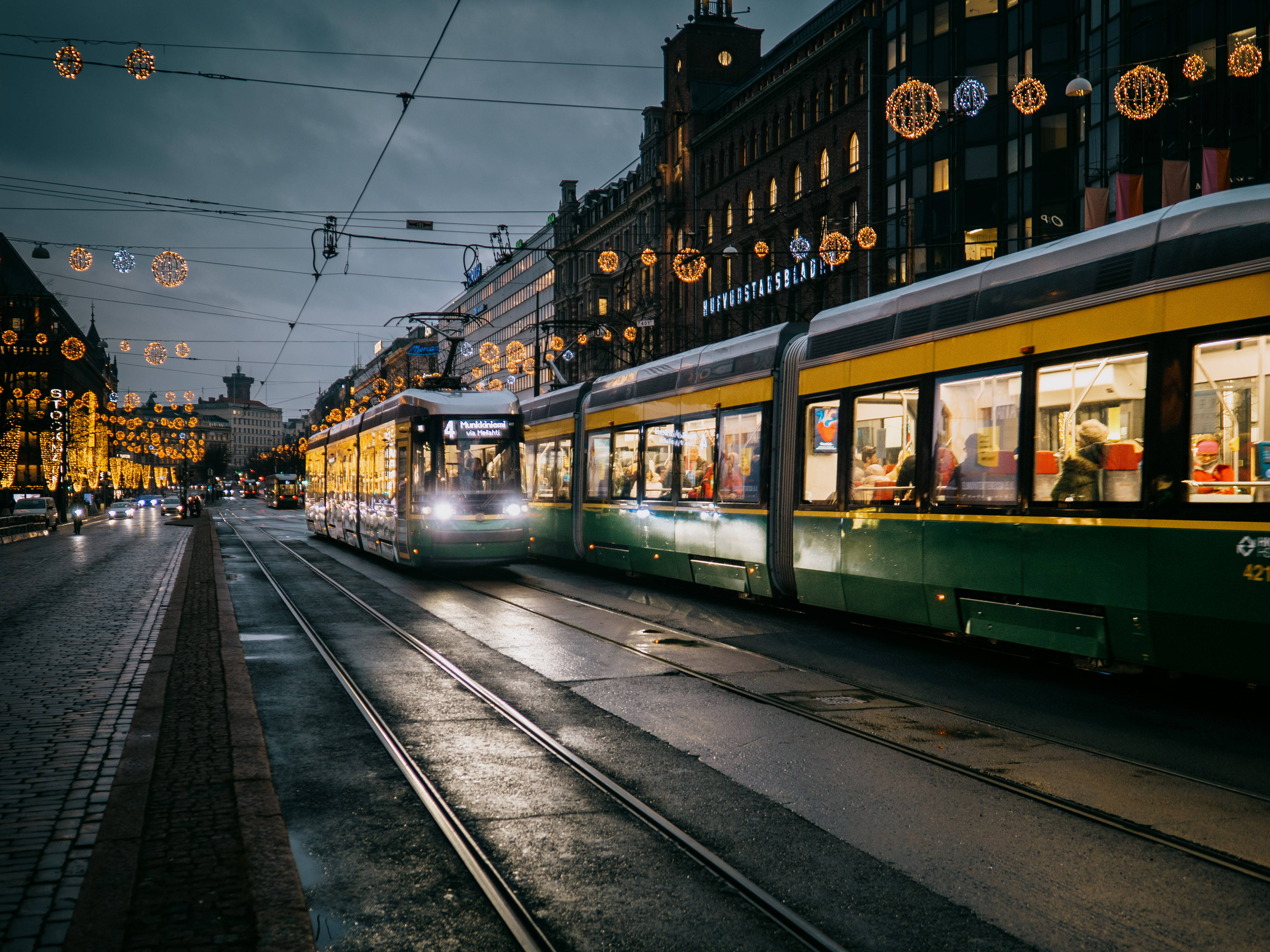 Finland 桌布 壁紙 — 一輛綠色和黃色的電車在黃昏時分滑過濕漉漉的城市街道，被節日燈串和發光的窗戶照亮。