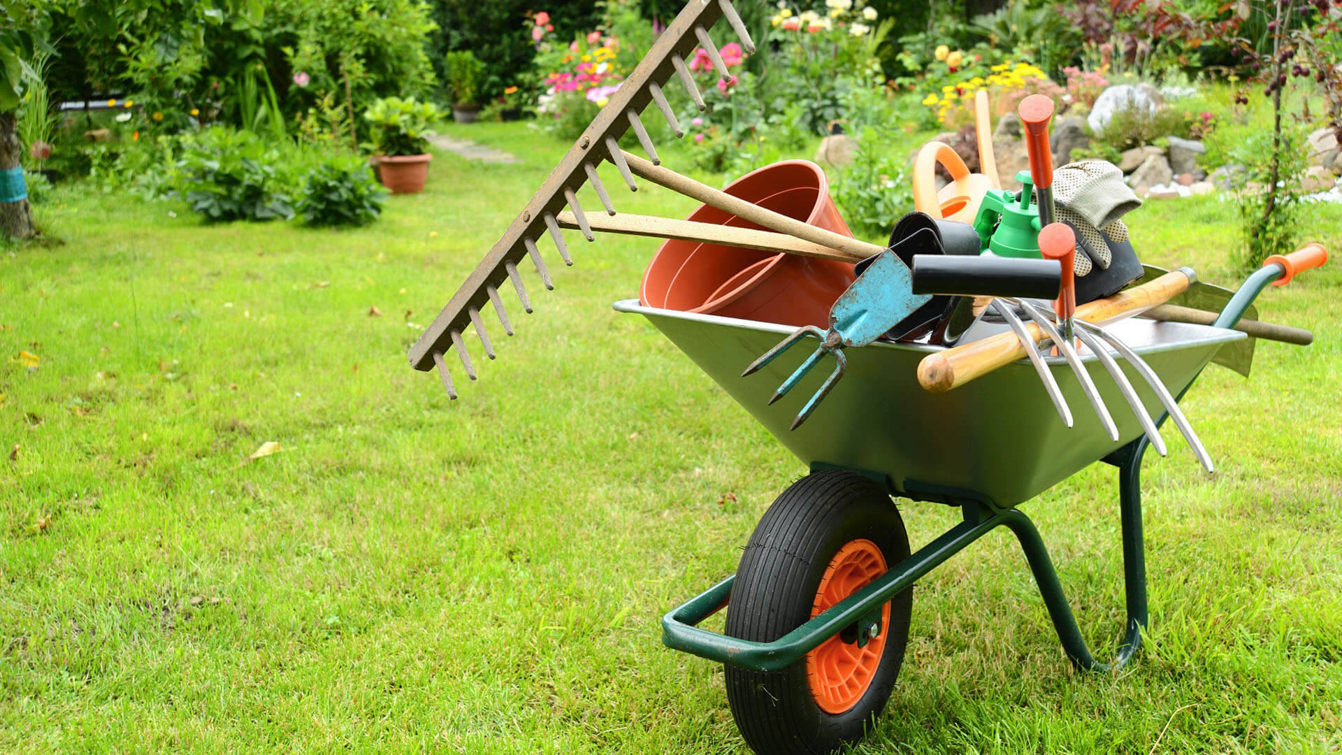 Gardening 桌布 壁紙 — 一輛綠色手推車裝滿園藝工具，放在綠油油的草地上，周圍有盛開的花朵和盆栽植物。