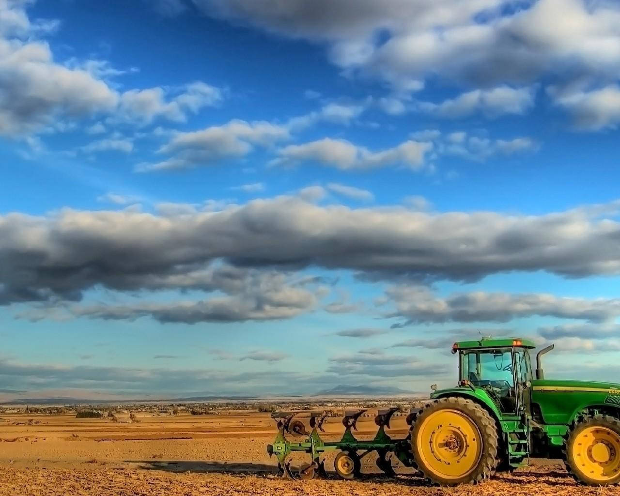 Agriculture 桌布 壁紙 — 一輛綠色約翰迪爾拖拉機帶黃色輪子，矗立在廣闊乾燥的田野中，上方是充滿蓬鬆白雲的戲劇性藍天。