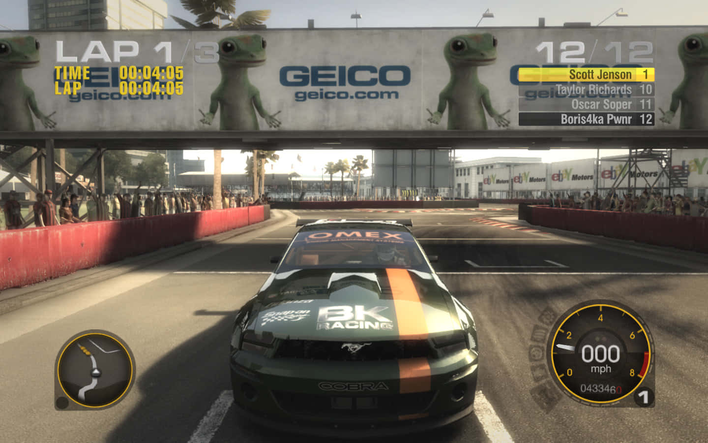 Grid 2 桌布 壁紙 — 一輛綠色賽車在陽光普照的賽道上疾馳，賽道上方有Geico廣告橫幅，上面有卡通青蛙。