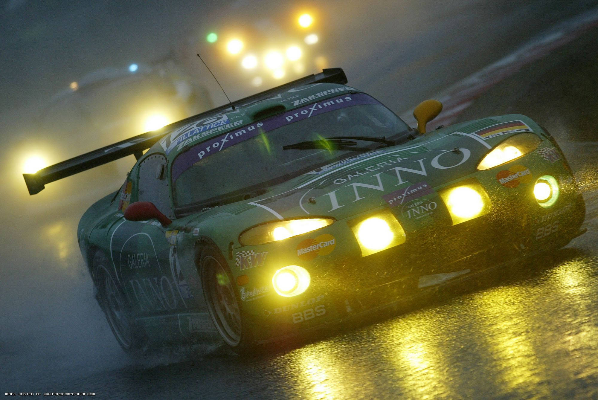 Racing 桌布 壁紙 — 一輛綠色賽車帶著發光的車頭燈在雨濕的夜間賽道上疾馳，背景有模糊的燈光。