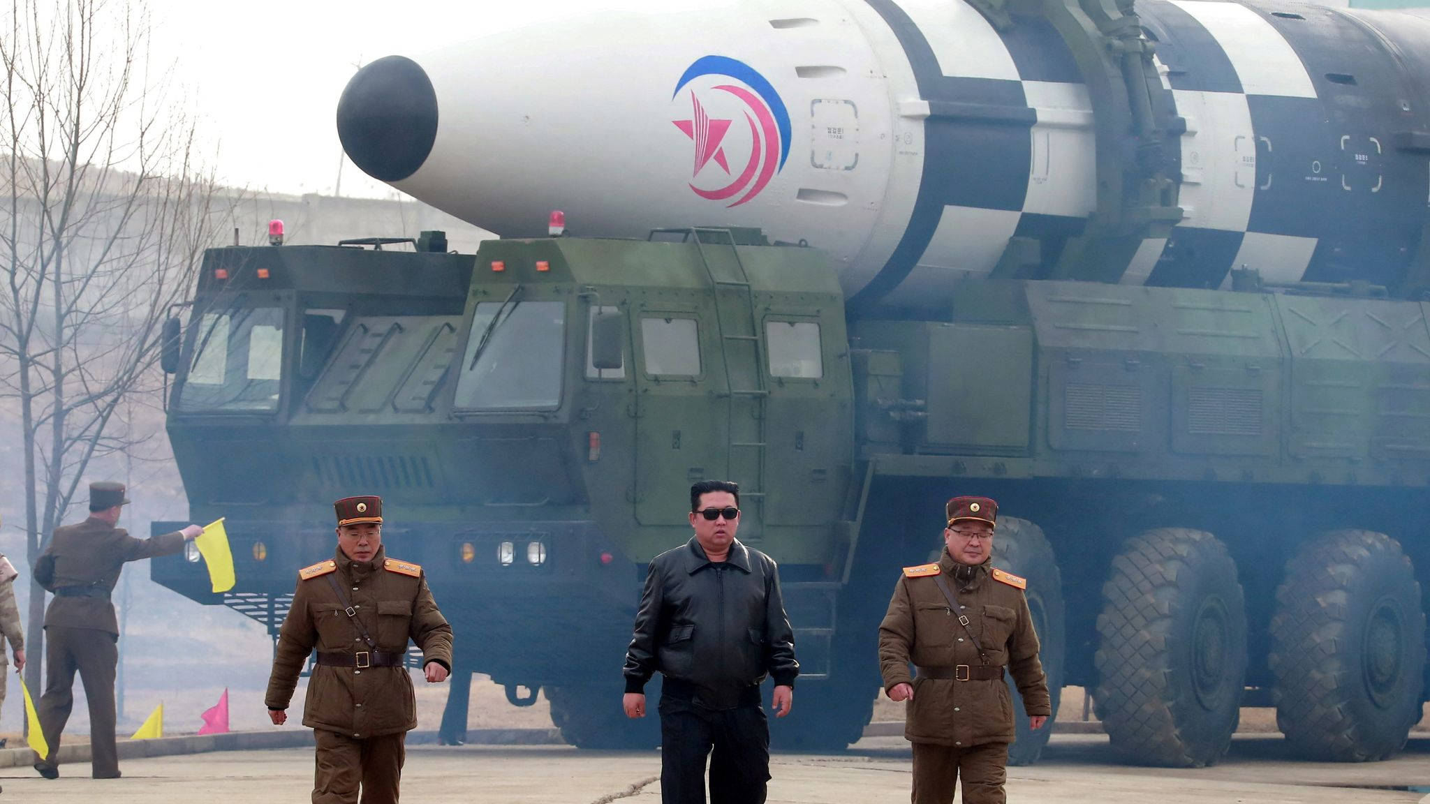 North Korea 桌布 壁紙 — 一輛綠色軍車上裝載著大型飛彈，身穿制服的官員在陰天中走在車前。