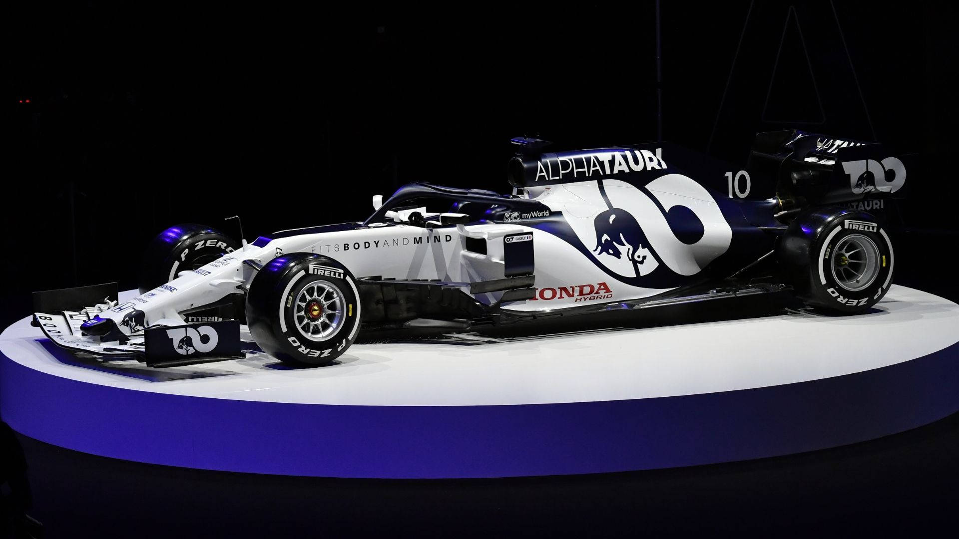 F1 桌布 壁紙 — 一輛線條流暢的AlphaTauri Honda一級方程式賽車，採用藍白配色，在戲劇性燈光下於圓形舞台上展示。