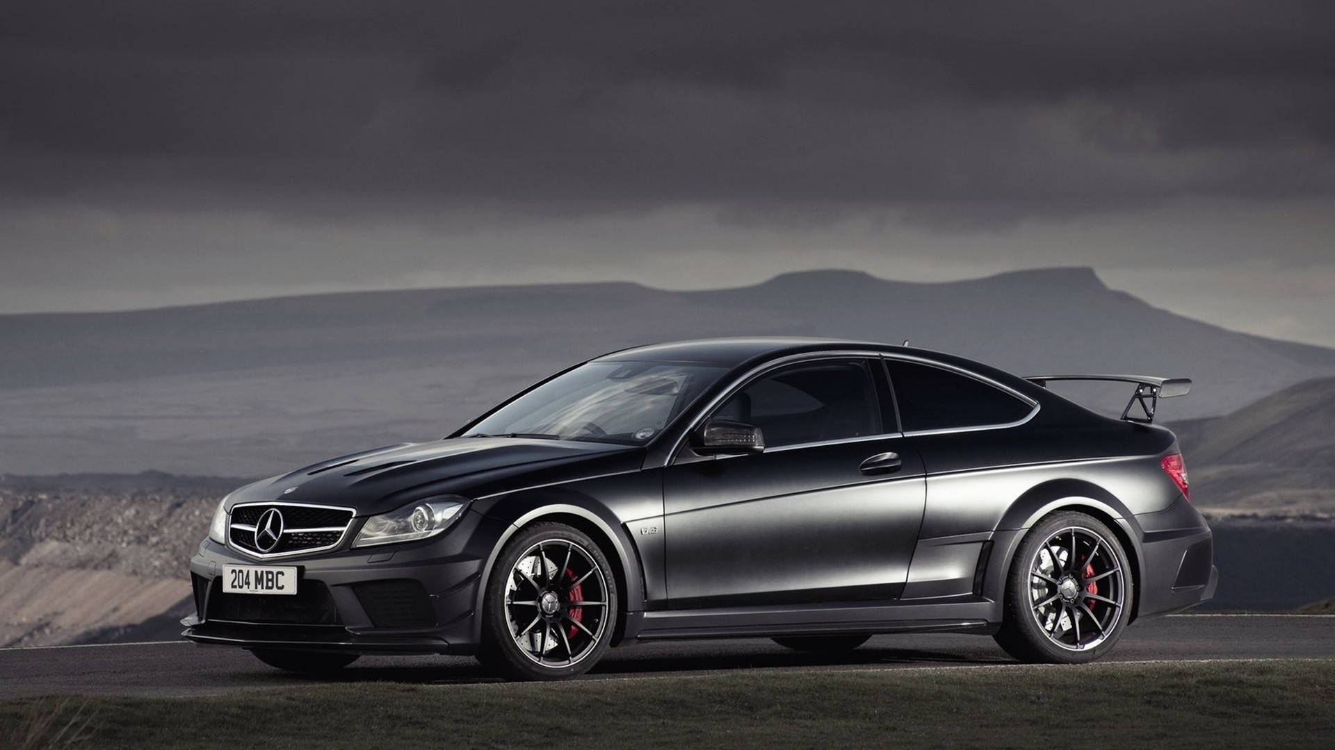 Mercedes Benz Amg 桌布 壁紙 — 一輛線條流暢的消光黑色賓士C63 AMG雙門轎跑車，配備紅色制動卡鉗，在戲劇性的多雲山景背景下停泊在道路上。