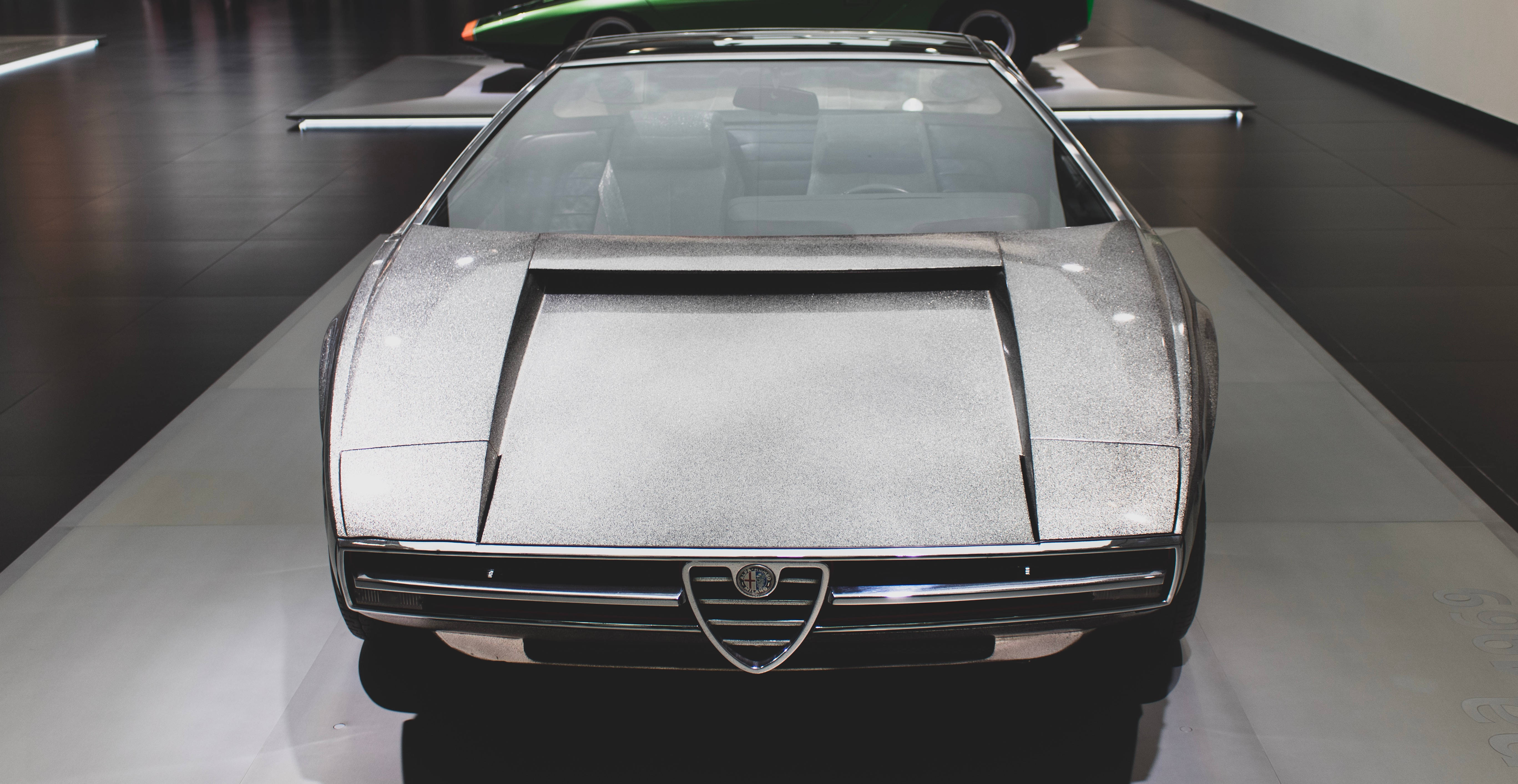 愛快羅密歐 桌布 壁紙 — 一輛線條流暢的銀色 Alfa Romeo 概念車，帶有閃閃發光的金屬飾面，在現代博物館環境中的白色平台上展示。