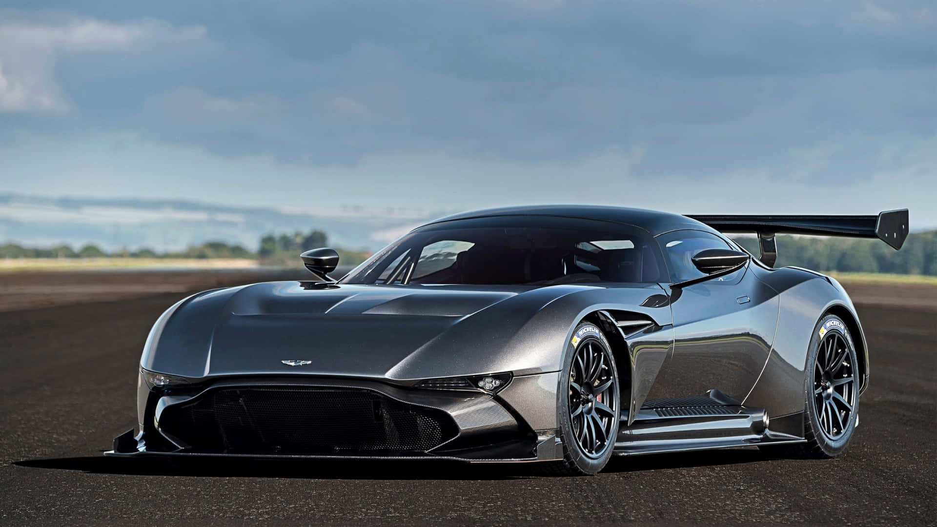 奧斯頓馬丁 桌布 壁紙 — 一輛線條流暢的銀色 Aston Martin Valkyrie 超級跑車，配備大型尾翼，在陰天停泊在瀝青賽道上。