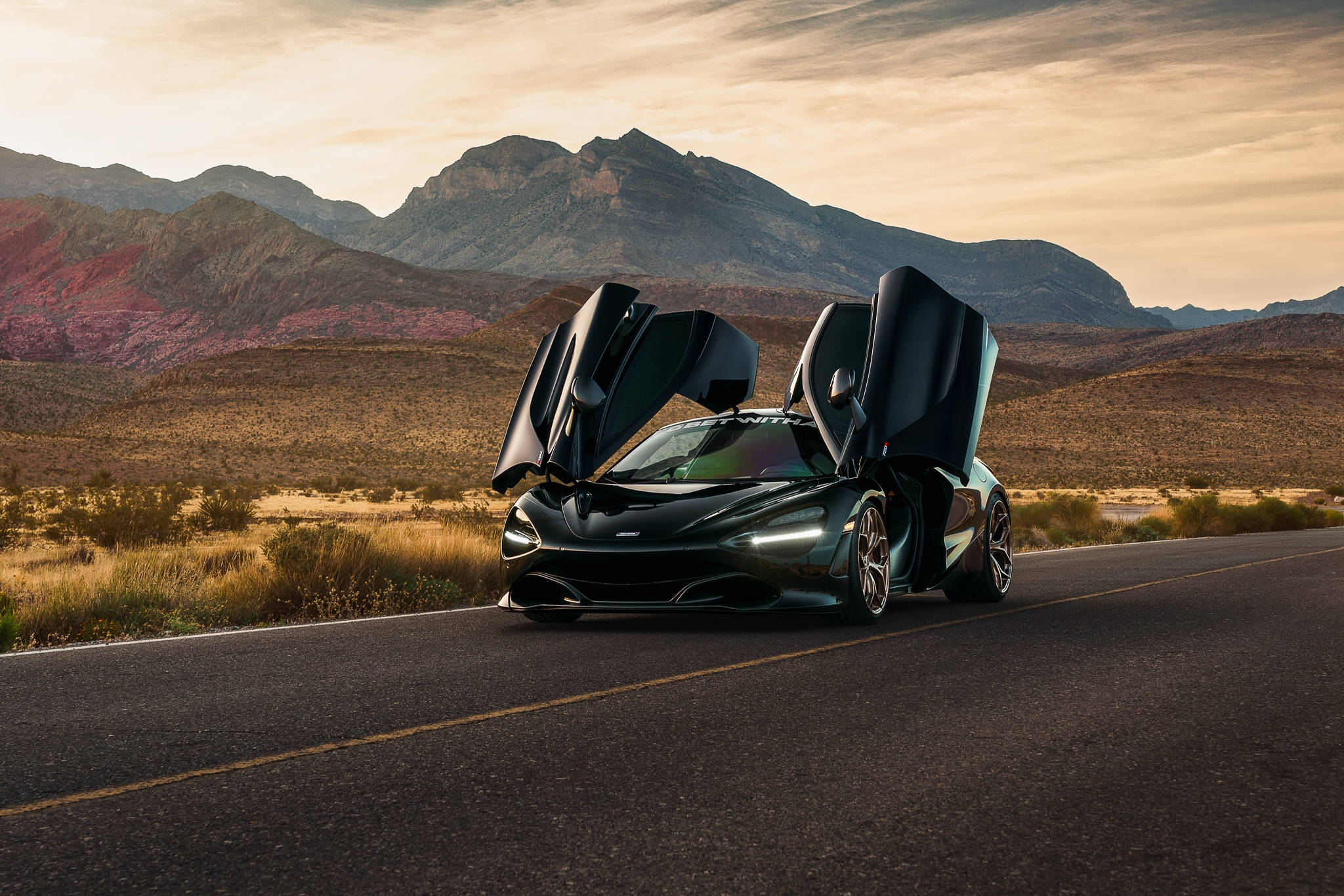 Mclaren 720s 桌布 壁紙 — 一輛線條流暢的黑色McLaren跑車，鷗翼車門敞開，停在沙漠公路上，背景是崎嶇的山脈，溫暖的夕陽映照天空。