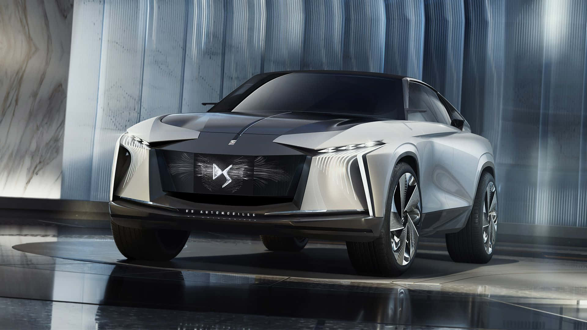 Ds Automobiles 桌布 壁紙 — 一輛線條銳利、車頭燈發光的時尚銀色未來電動SUV，展示在配備反光地板和金屬牆壁的高科技展廳中。