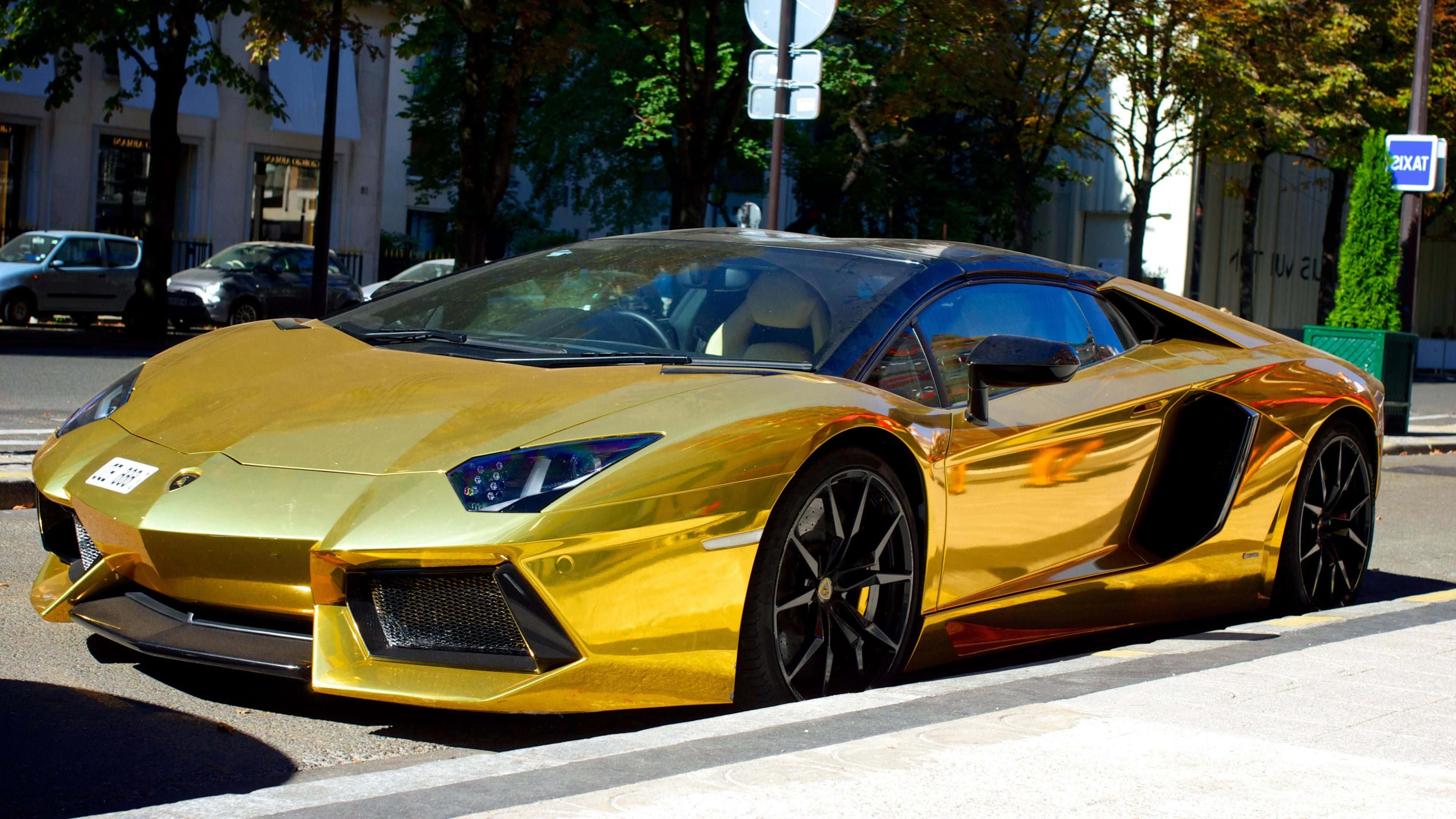 跑車 桌布 壁紙 — 一輛耀眼的金色藍寶堅尼Aventador停泊在綠樹成蔭的城市街道上，反射著陽光。