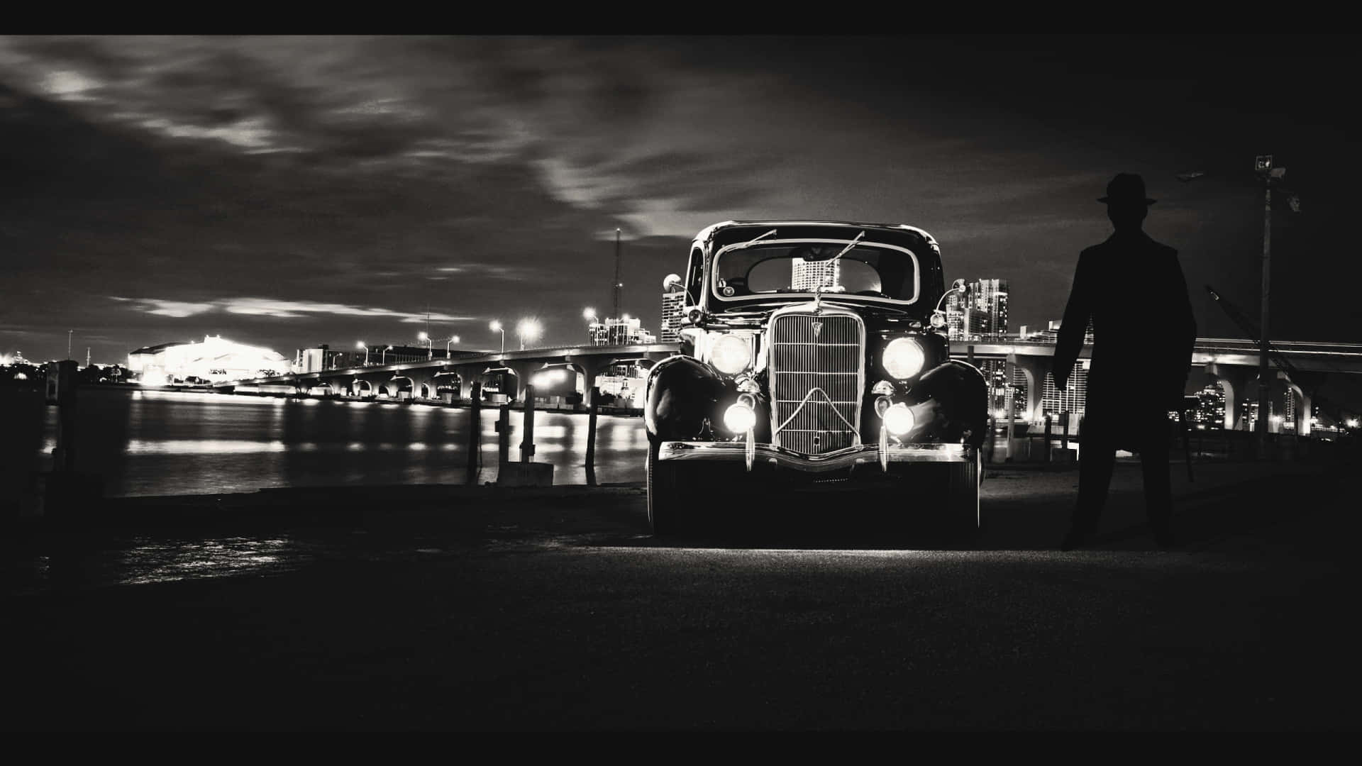 Classic Movie 桌布 壁紙 — 一輛老爺車的車頭燈劃破黑夜，一個剪影男子站在它旁邊，靠近海濱橋樑。